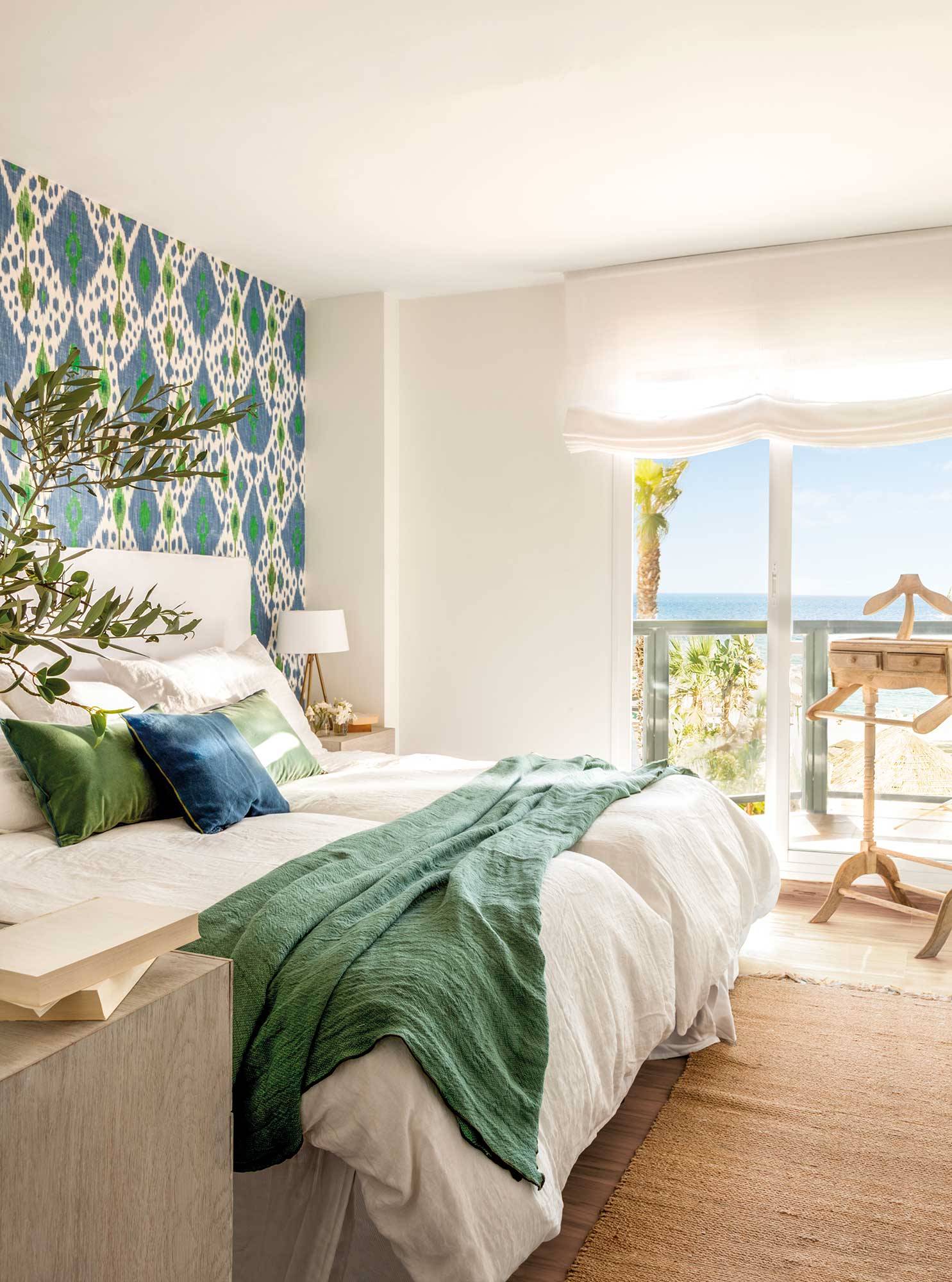 Dormitorio blanco con pared del cabecero con papel pintado con motivos geométricos verdes y azules.