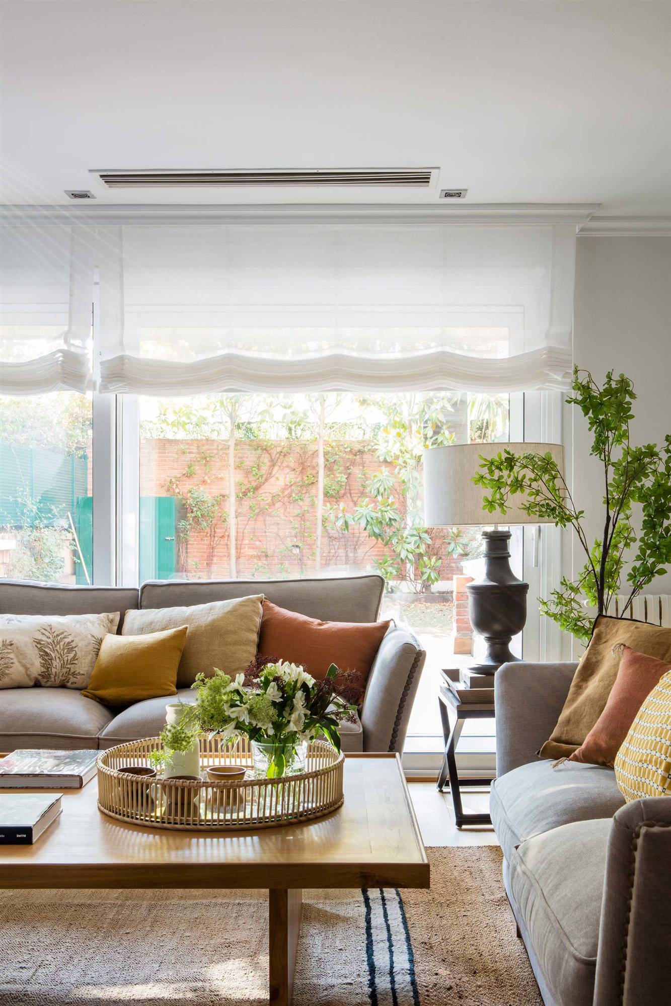 Salón con dos sofás de color gris dispuestos en forma de L y amplios ventanales con estores. 