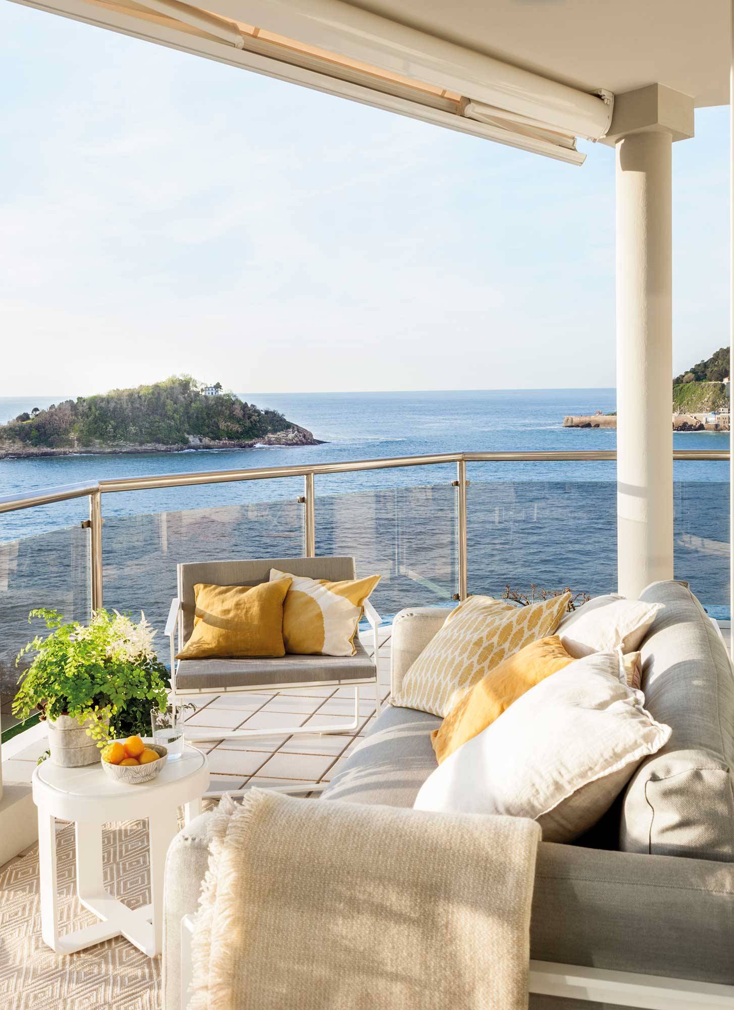 Un balcón mirando al mar con sofá, butaca y plaid. 