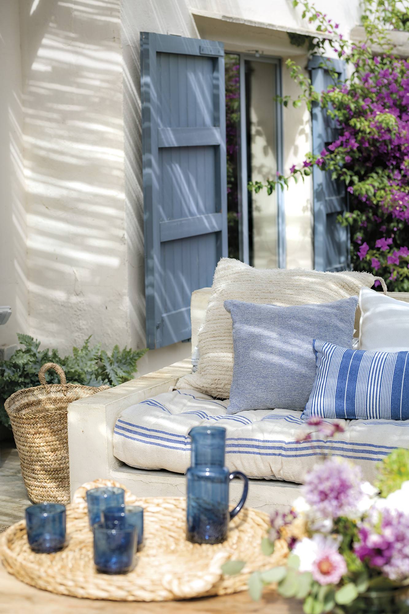 Porche de estilo mediterráneo con sofá de obra con cojines azules y mesa de centro de madera con bandeja de fibras.