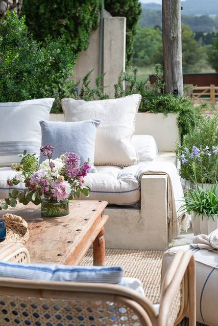 Porche con sofá de obra, cojines en tonos azules, mesa de centro de madera con un jarrón con flores y plantas de exterior.