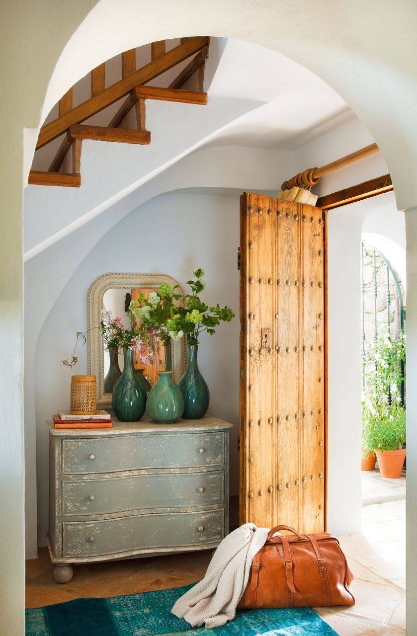 Recibidor con puerta de entrada de madera y cómoda con cajones restauradas con jarrones verdes y alfombra sobre suelo parquet.