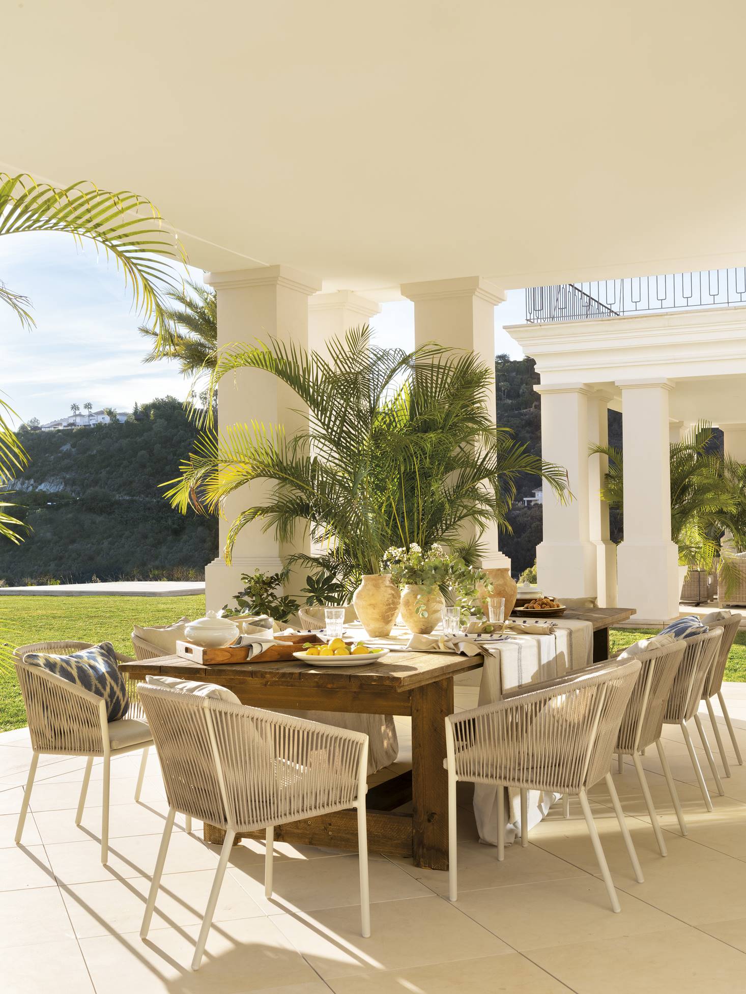 Comedor con mesa de madera y sillas blancas en un porche con vistas al jardín.