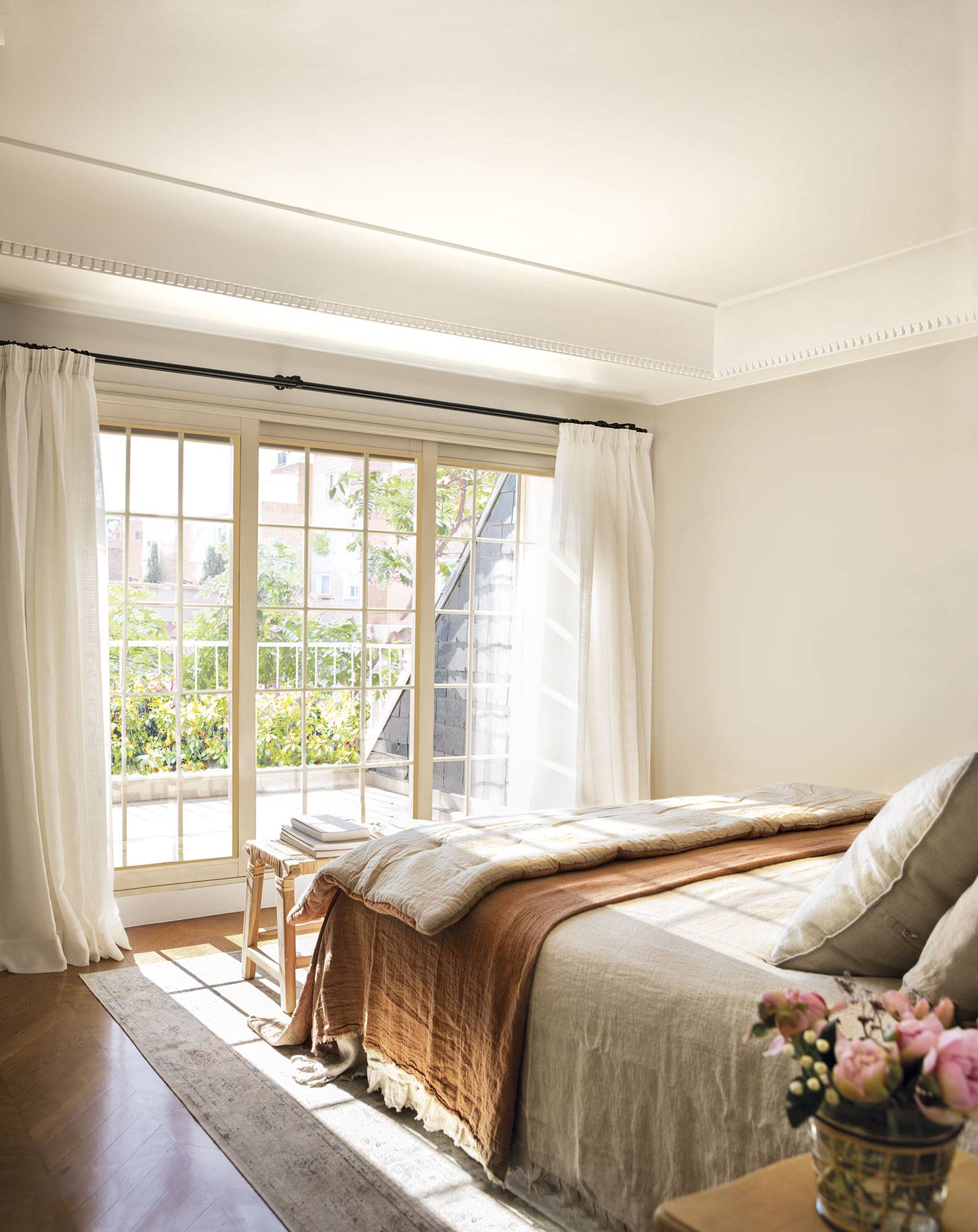 Dormitorio en suite con cama en tonos tierra con plaid, banqueta y ventanal con cortinas de lino en blanco roto. 