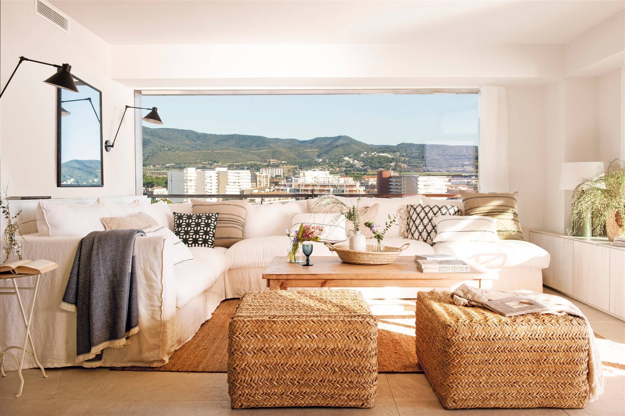 Salón con sofá con funda blanca, pufs y alfombra de fibra natural y gran ventanal. 