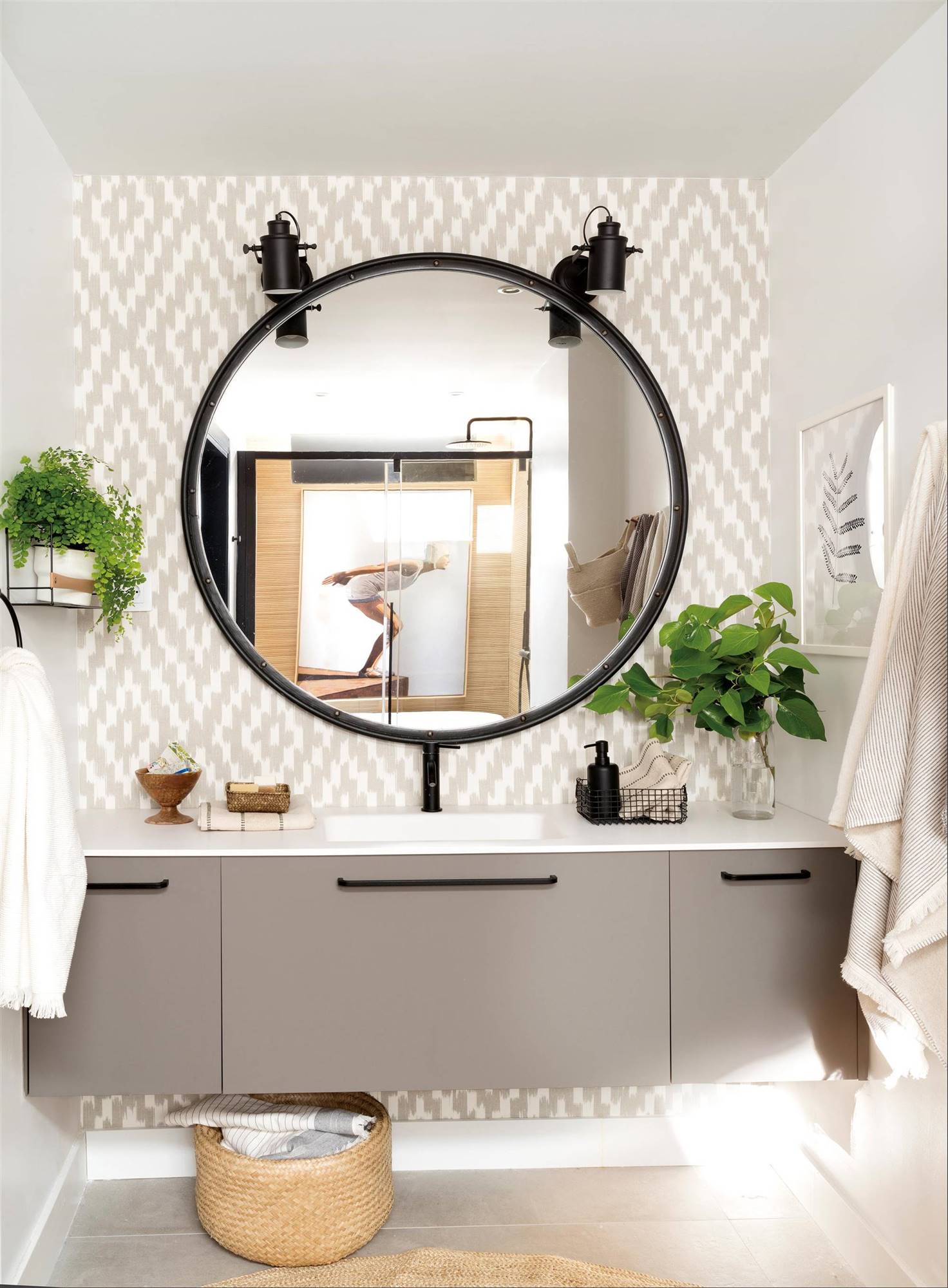 Baño con papel pintado e na pared, espejo redondo y mueble de color gris. 