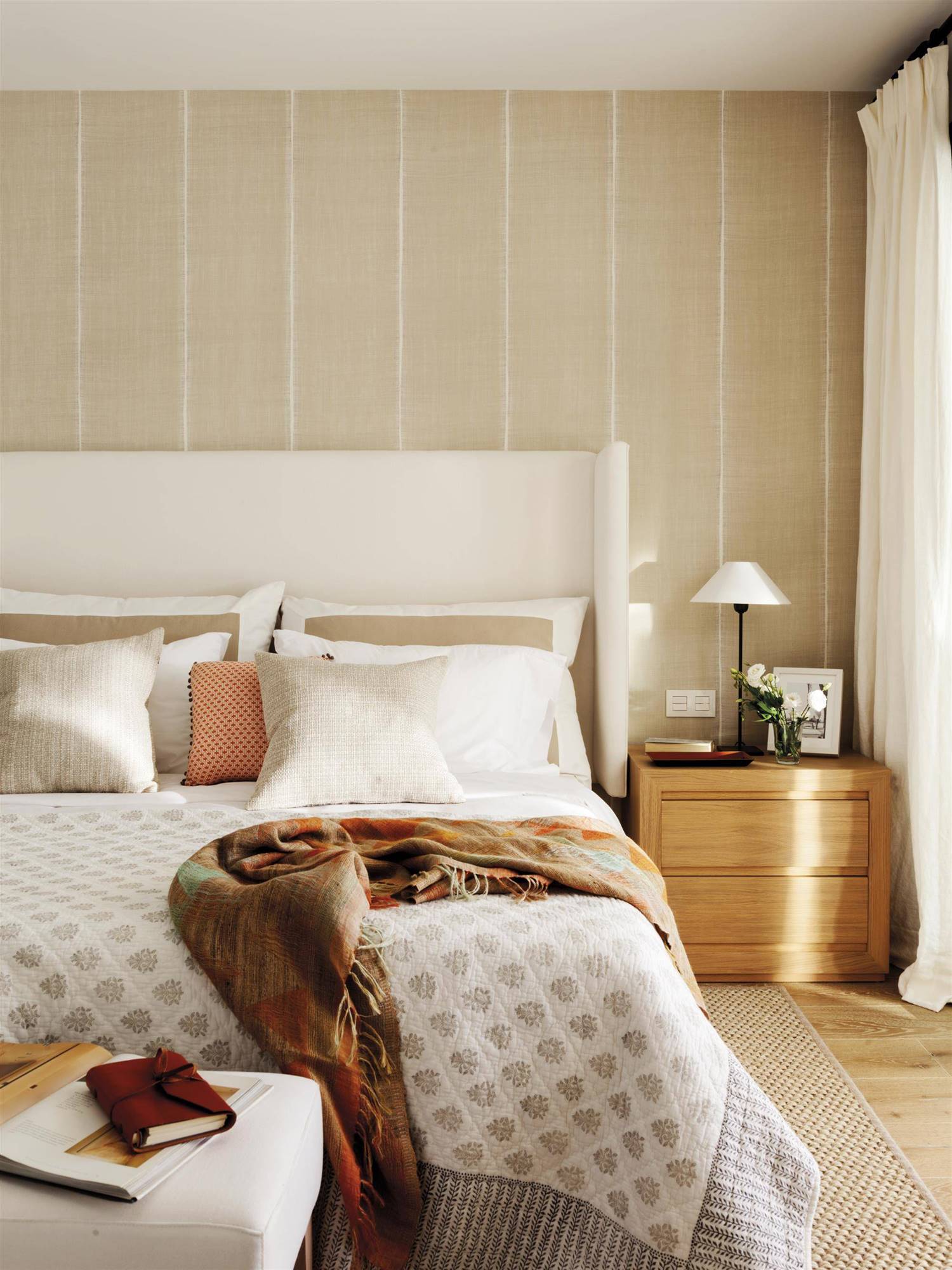 Dormitorio con cabecero orejero beige, mesita de noche de madera y papel pintado a rayas.