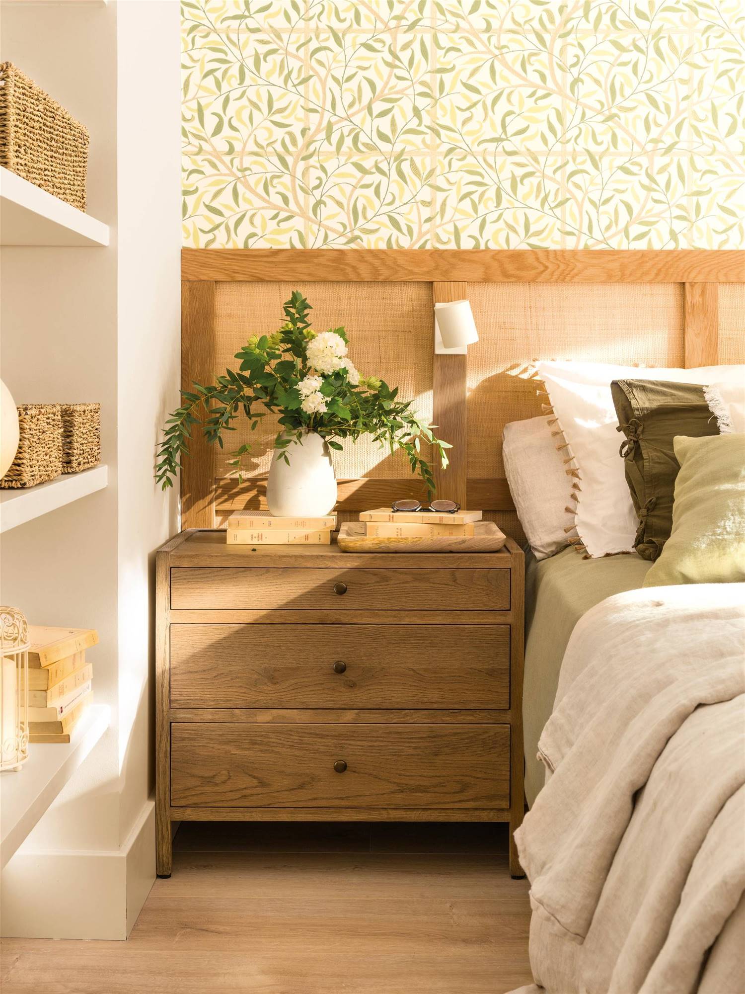 Dormitorio con cabecero de madera y rejilla sobre papel pintado con motivos botánicos y mesita de noche de madera.