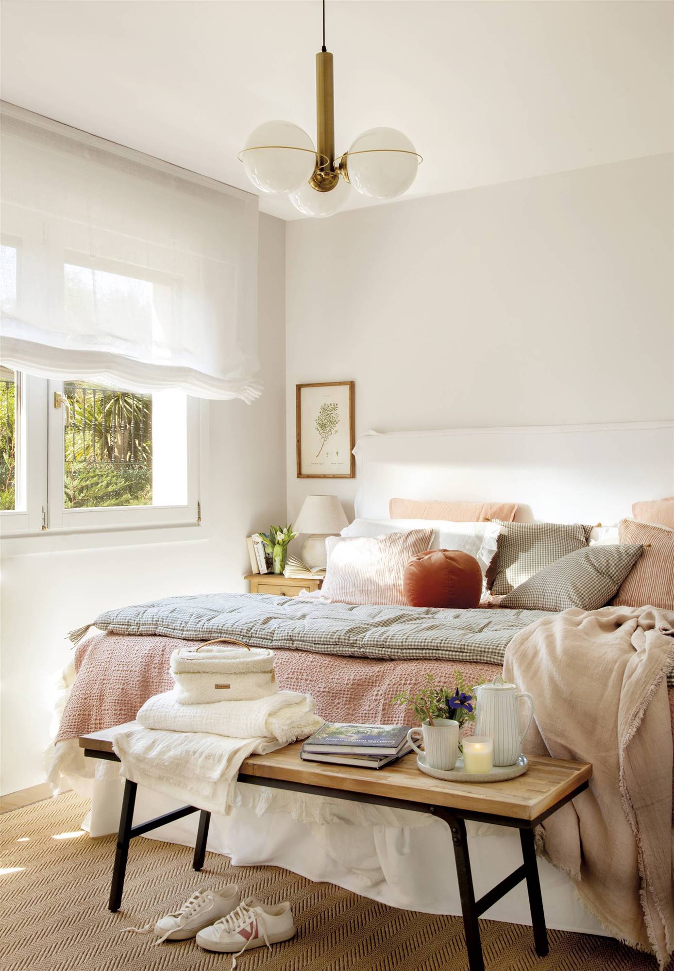 Dormitorio de color blanco, con cabecero y estores blancos, lámpara de techo con bolas, ropa de cama en tonos rosados y banco a los pies. 