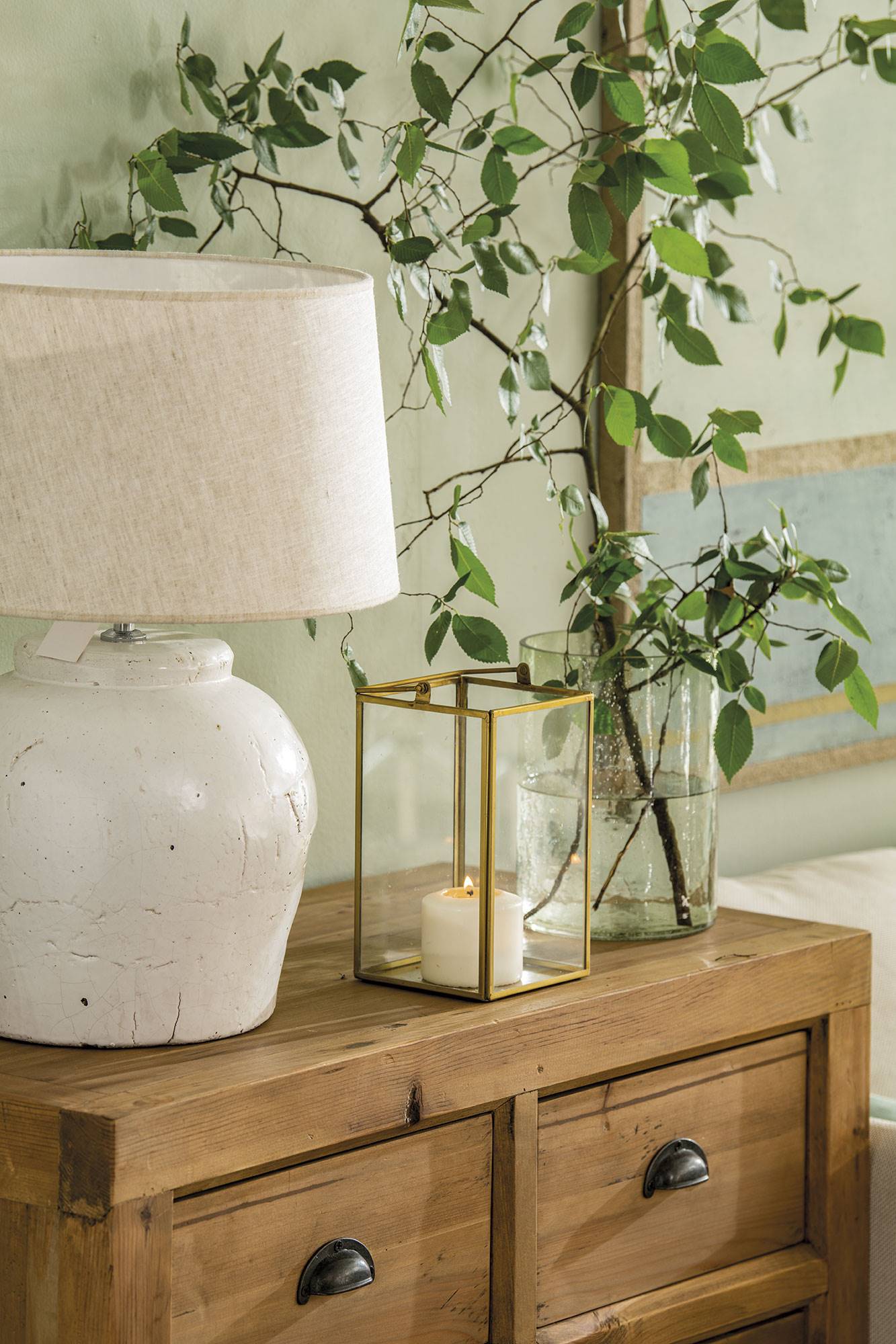 Cómoda de madera con lámpara de mesa de cerámica blanca, candelabro de cristal y latón con una vela y jarrón.