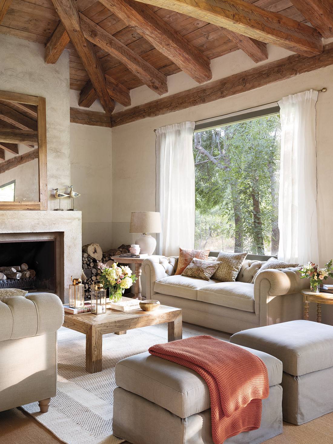  Salón con chimenea y techo de madera con sofás de capitoné, pufs con faldón y alfombra de cenefa contrastada.