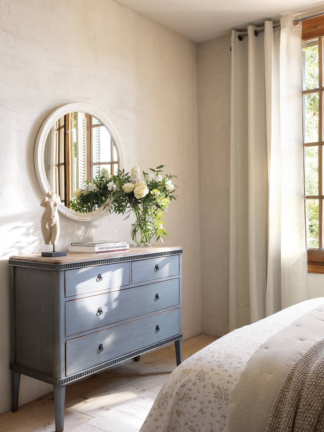  Dormitorio con cómoda con cajones azul, flores y espejo blanco redondo con moldura envejecida.