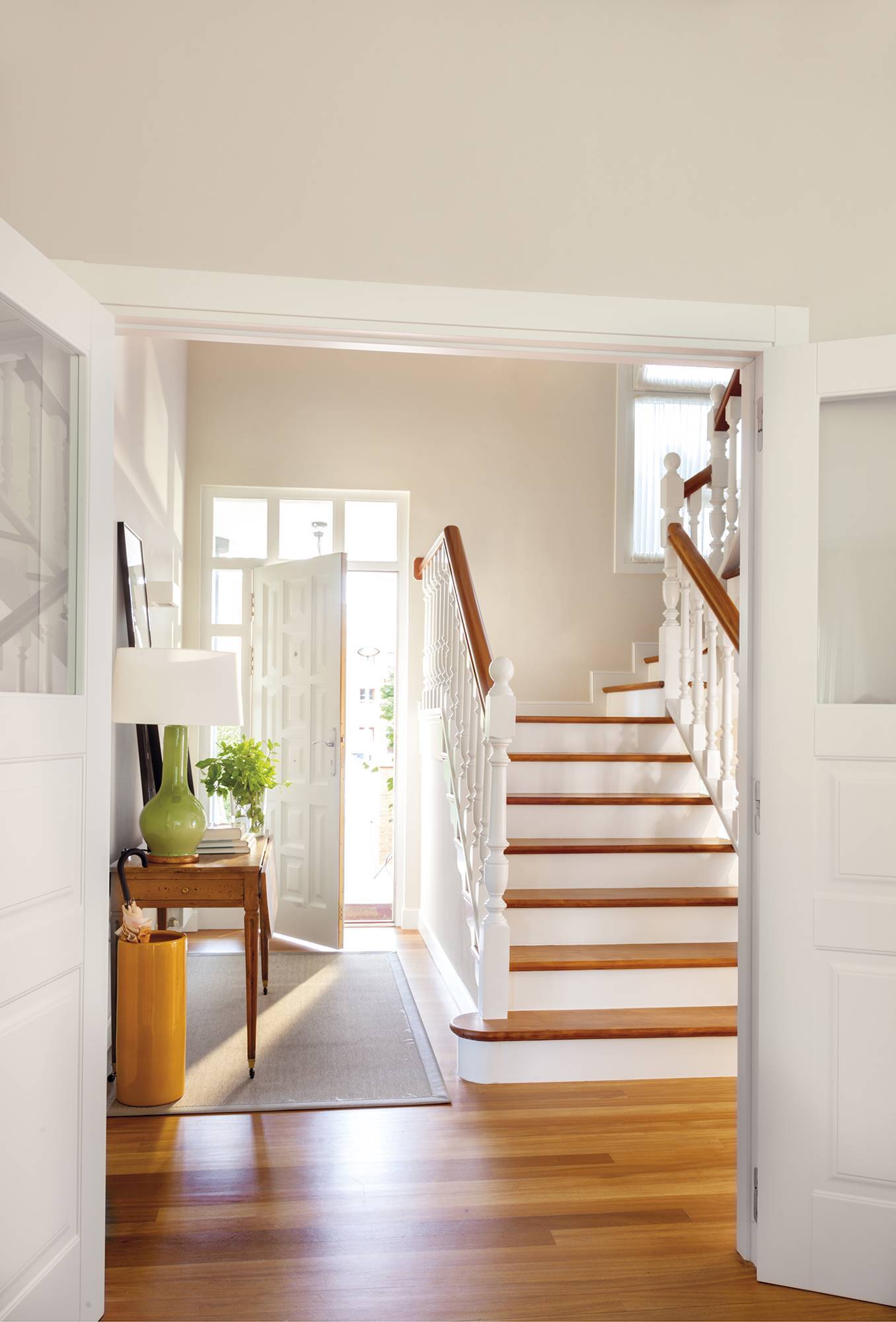 Recibidor luminoso con escalera de madera y barandilla pintada de blanco.