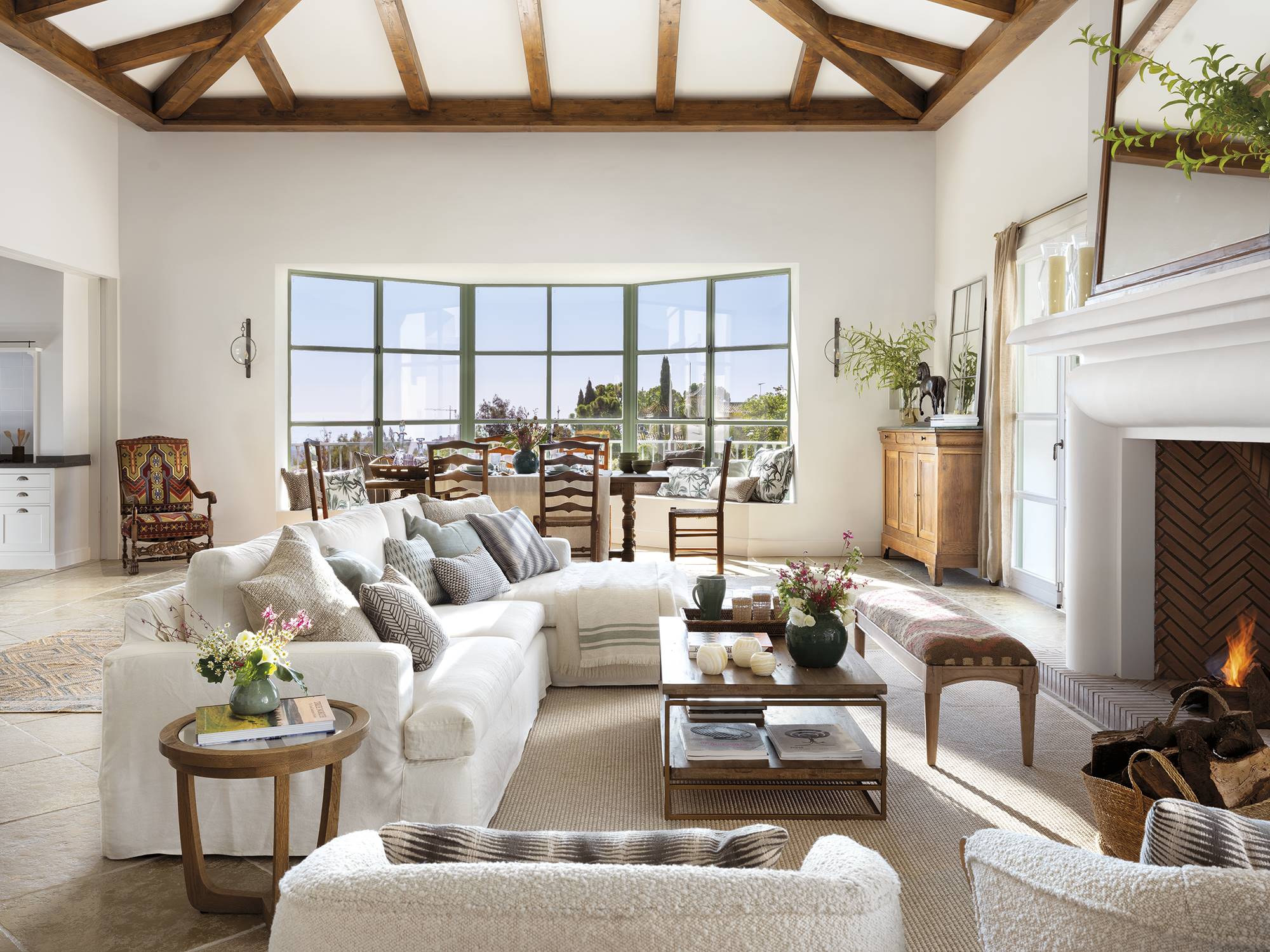 Salón clásico con vigas en el techo, ventanal verde con banco, chimenea, sofá con chaise longue blanco y alfombra.