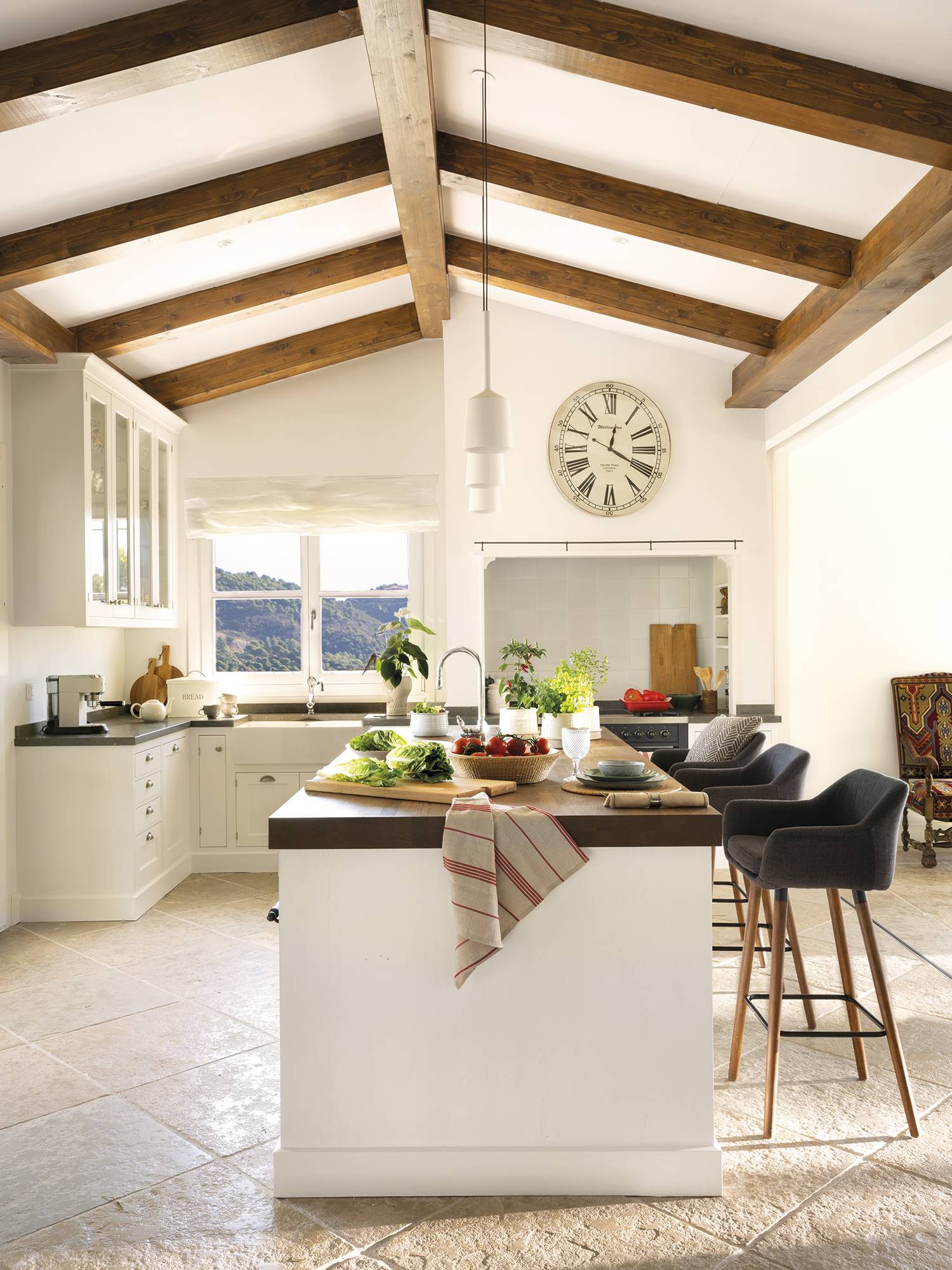 Cocina grande con isla, taburetes, mobiliario blanco y vigas de madera en el techo. 