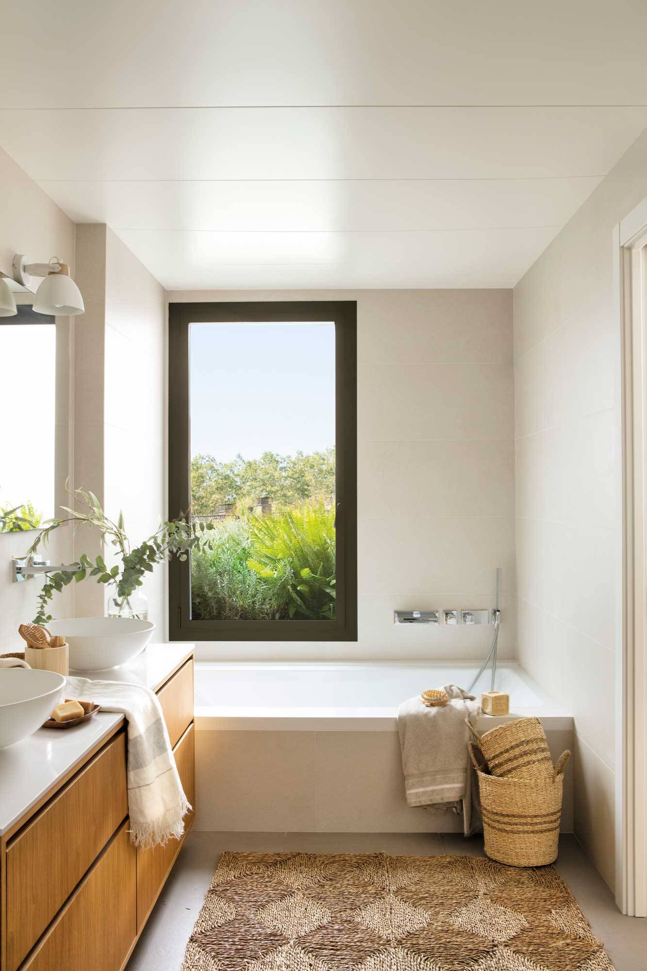 Baño con mueble de madera, bañera bajo el ventanal y alfombra de fibra.