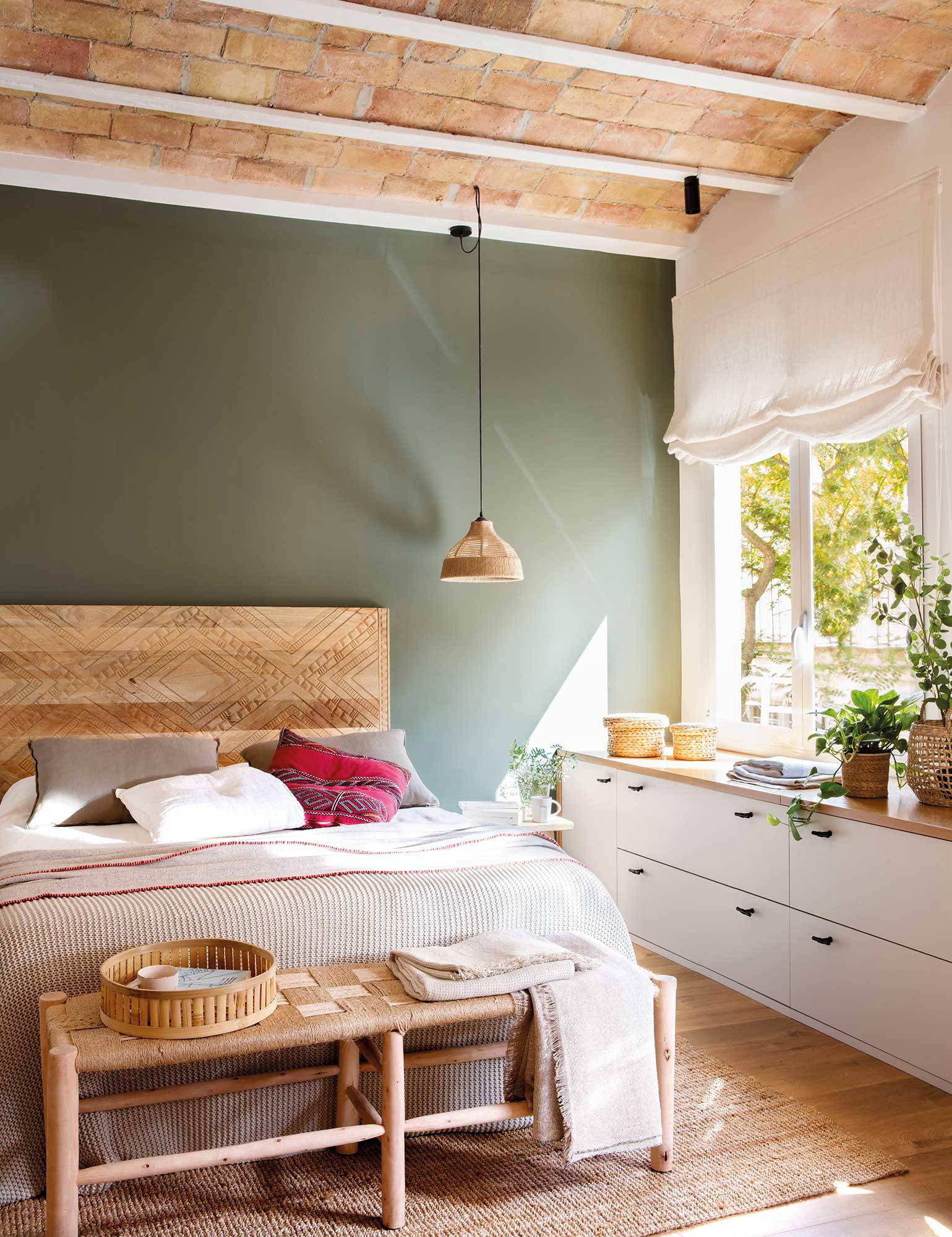 Dormitorio con pared verde, cabecero de madera y bajo de la ventana aprovechado con una cajonera. 
