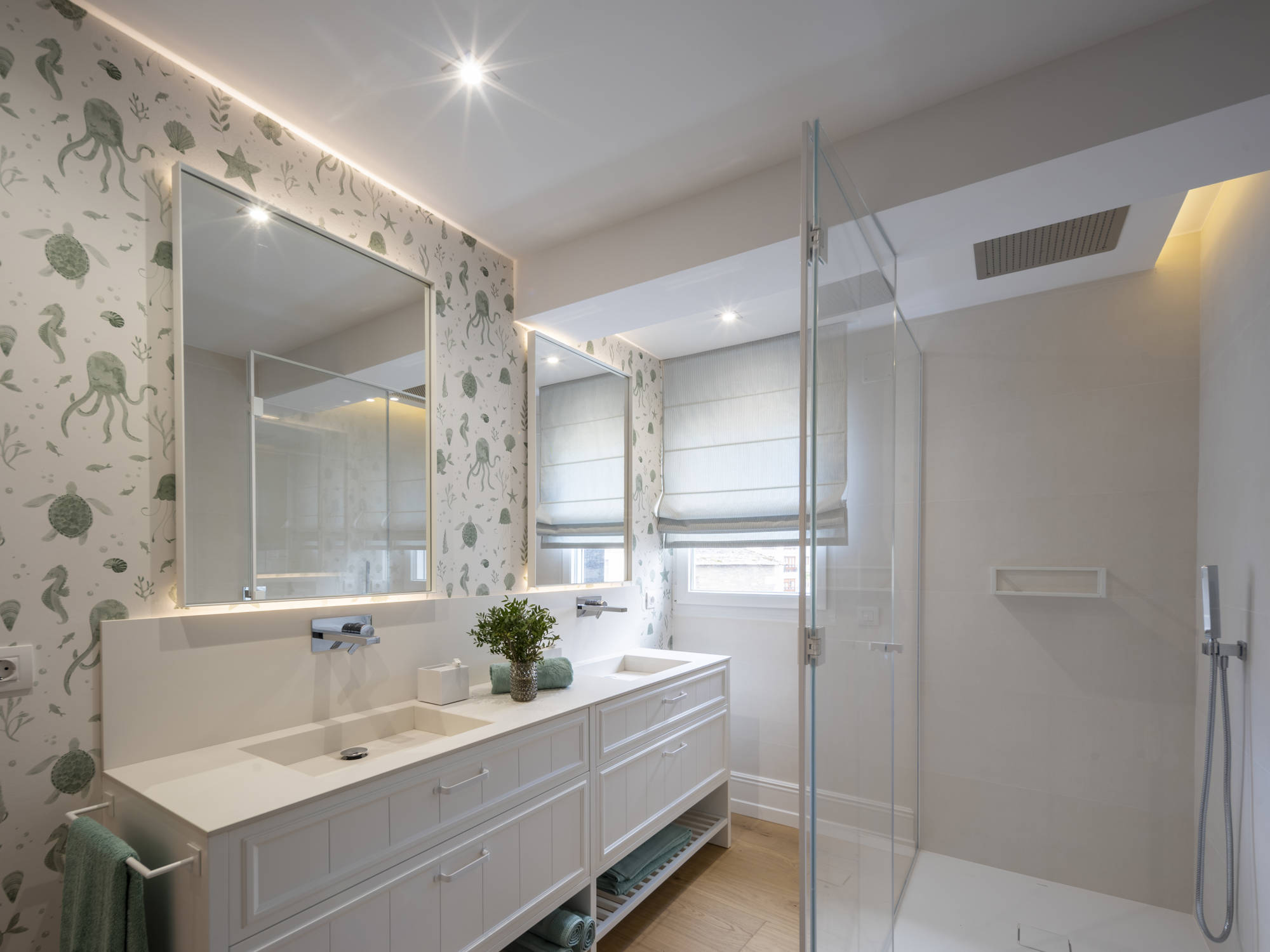 Baño con ducha con mampara de cristal, dos lavamanos y espejos iluminados, y papel pintado de temática marina.