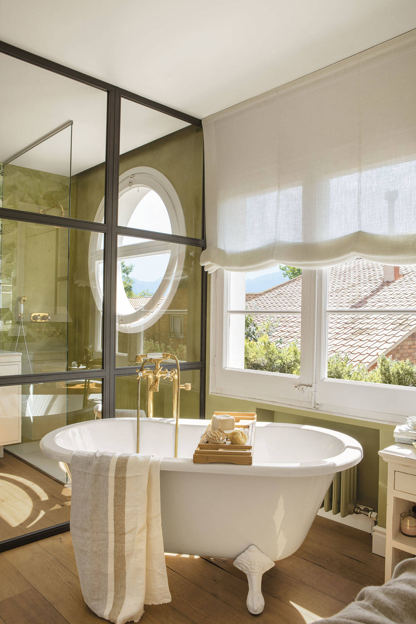 Baño con paredes verdes, ventana redonda, pared de cristal y bañera exenta.