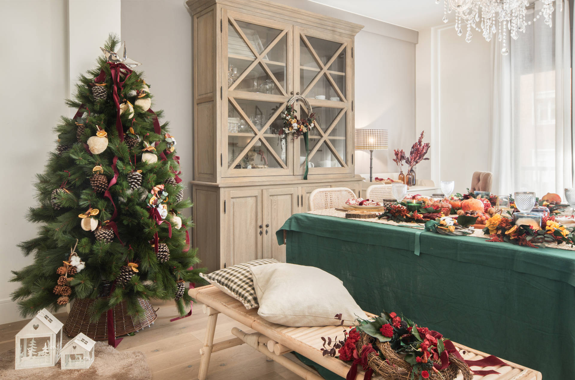Comedor navideño con árbol de Navidad decorado con piñas, gajos de naranja, estrella plateada y lazo rojo. Bajo el árbol, textil de cordero y casitas blancas de adorno. 