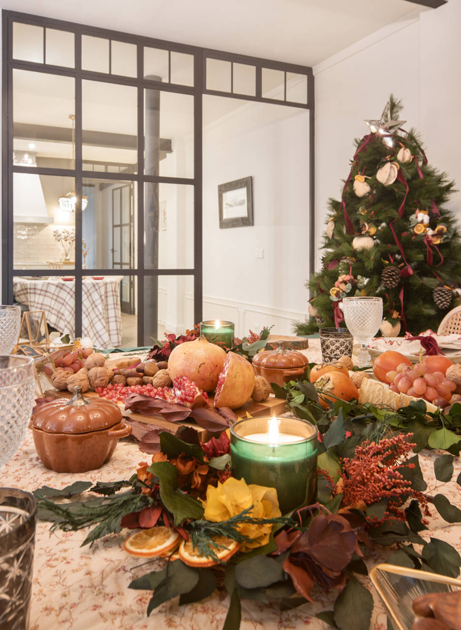 Mesa de Navidad con mantel navideño, velas, flores secas, vajilla de cristal, vegetales y tablas de madera. 