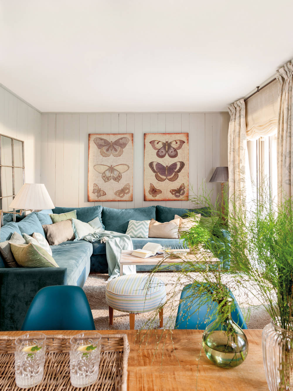 Salón con sofá verde rinconero, láminas con mariposas y mesa con plantas de interior.