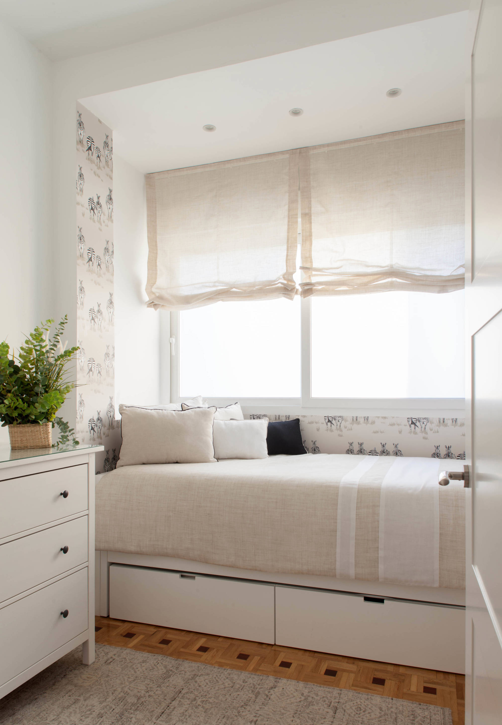 Dormitorio con papel pintado, estores, cama con espacio de almacenaje y cómoda blanca. 