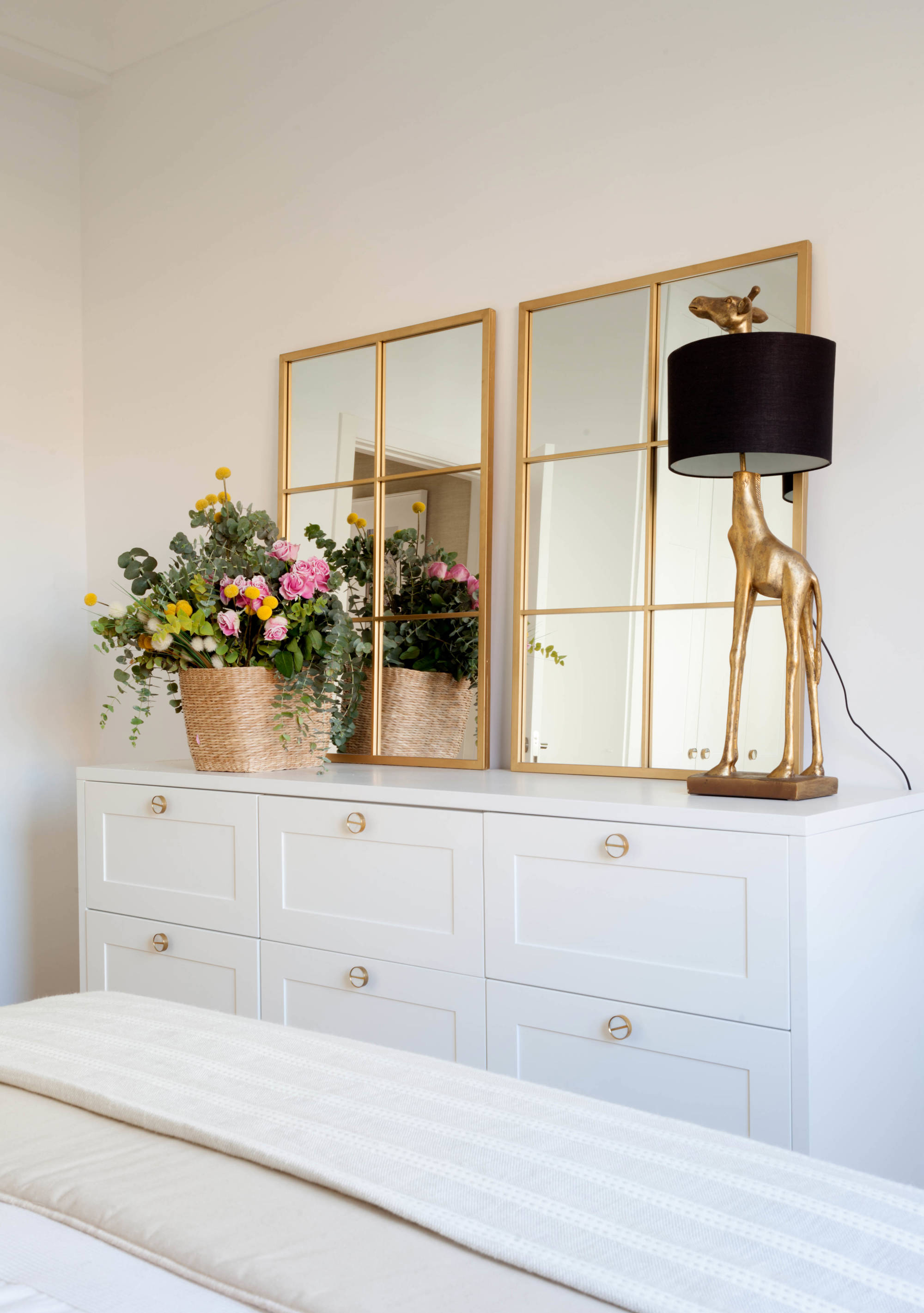 Dormitorio principal con cómoda blanca con espejos de cuarterones dorados, lámpara en forma de jirafa dorada y cesta con flores.