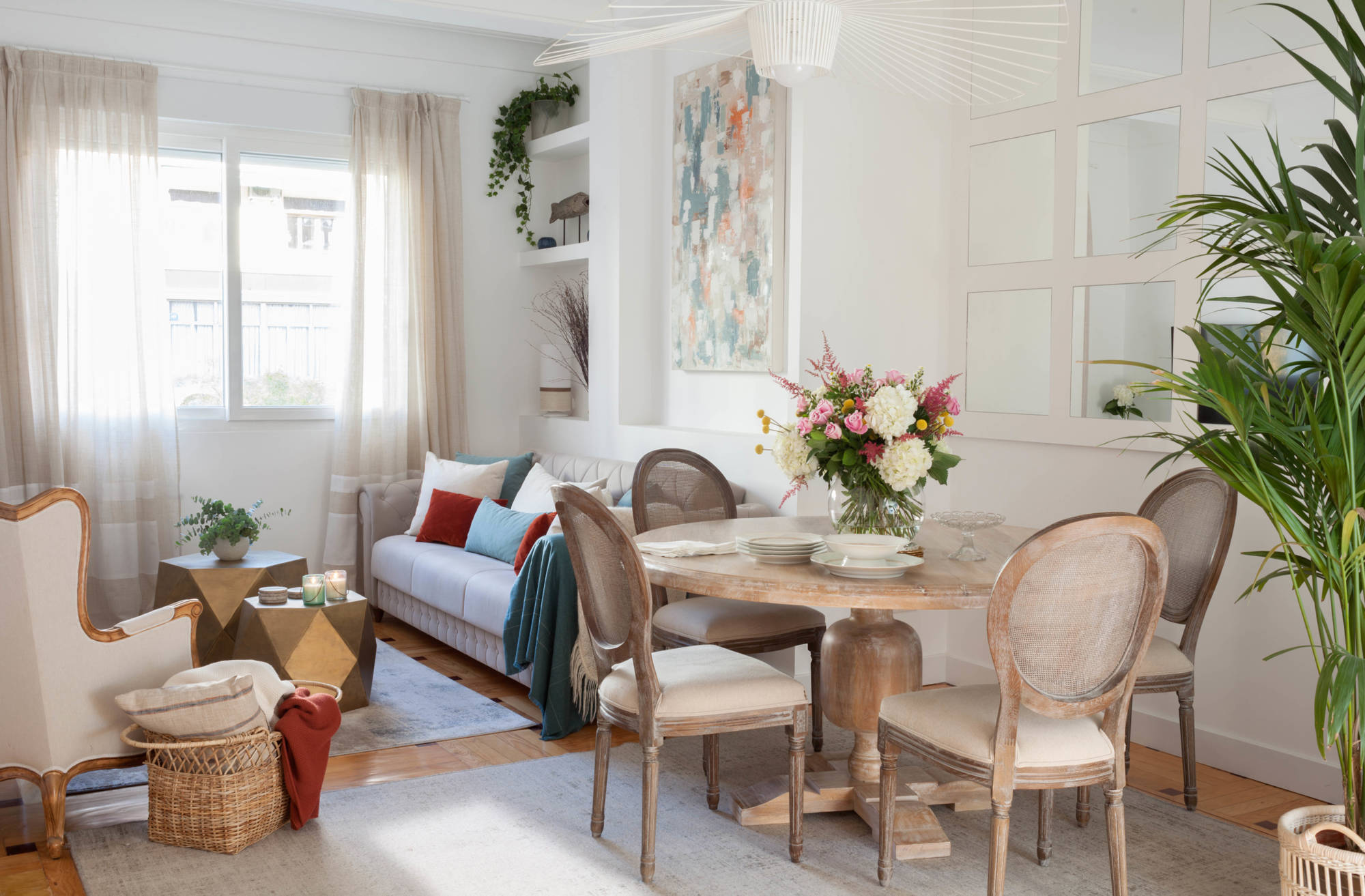 Salón comedor con sofá beige, alfombras grises, mesa de comedor de madera, espejo de cuarterones y mesas de centro doradas. 