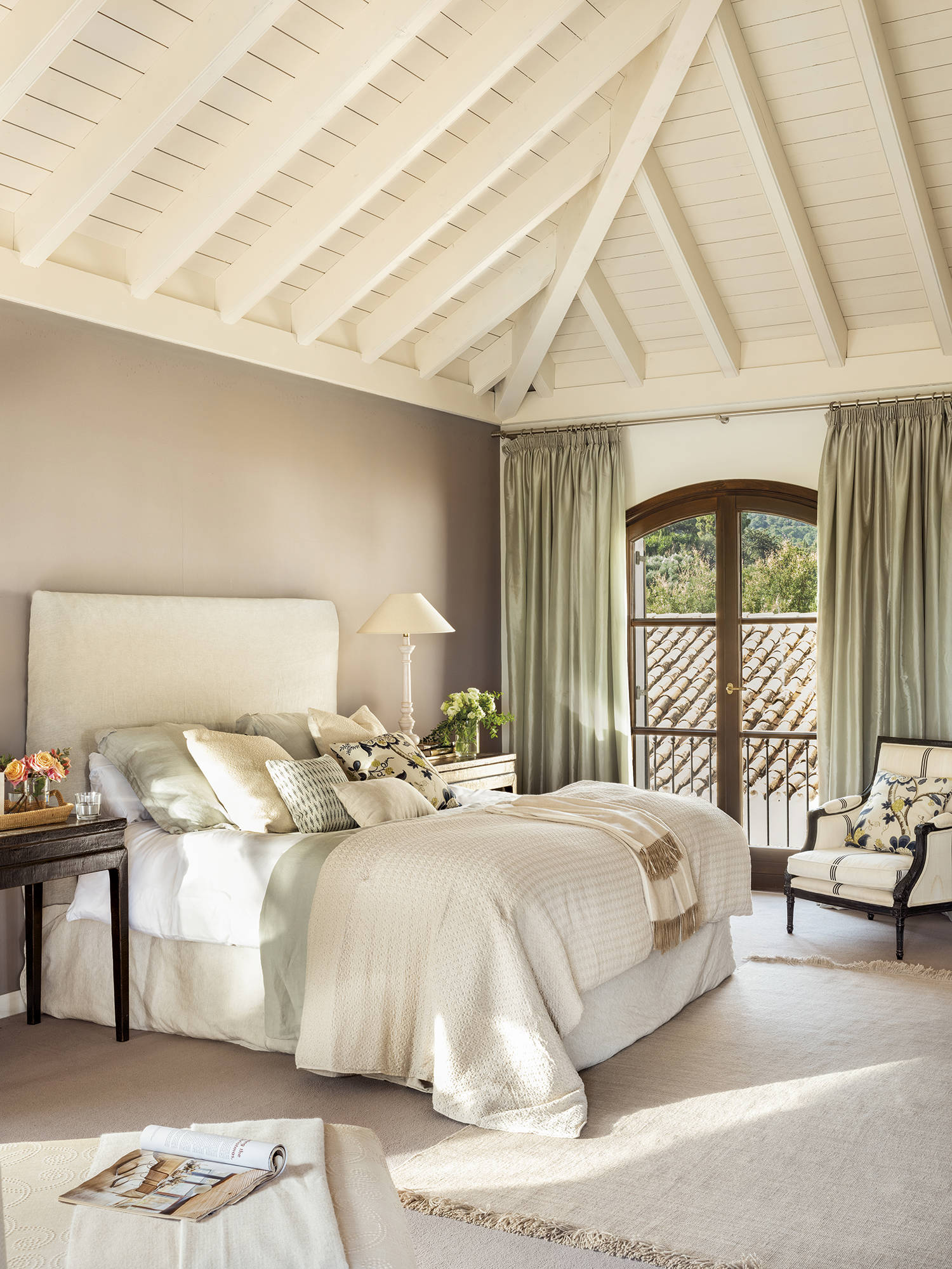 Dormitorio principal con cama, aflombra, butaca, cabecero blanco y techo abuhardillado con vigas pintadas de blanco