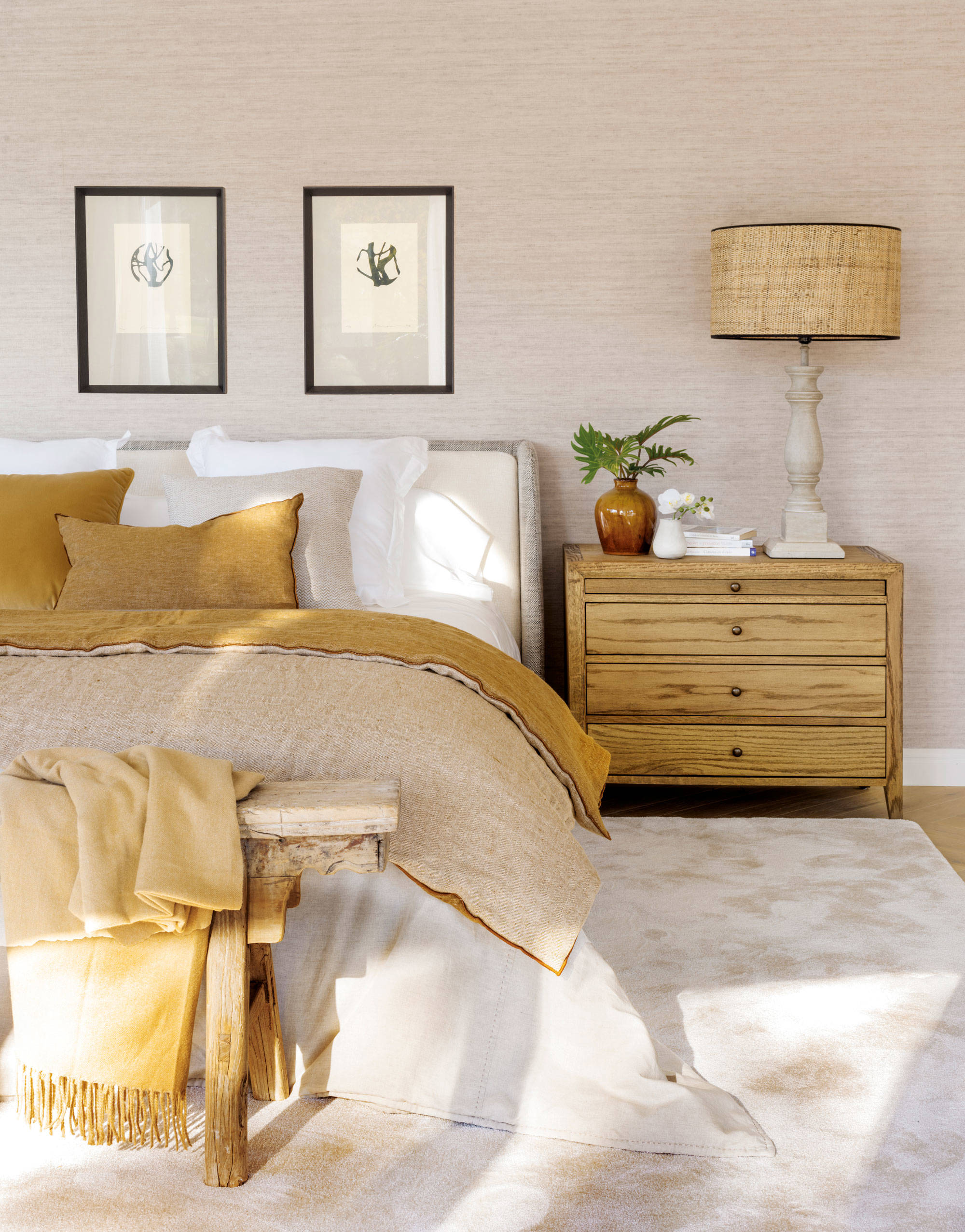 Dormitorio de primavera con mesilla de madera clara, cuadros y ropa de cama mostaza y gris.