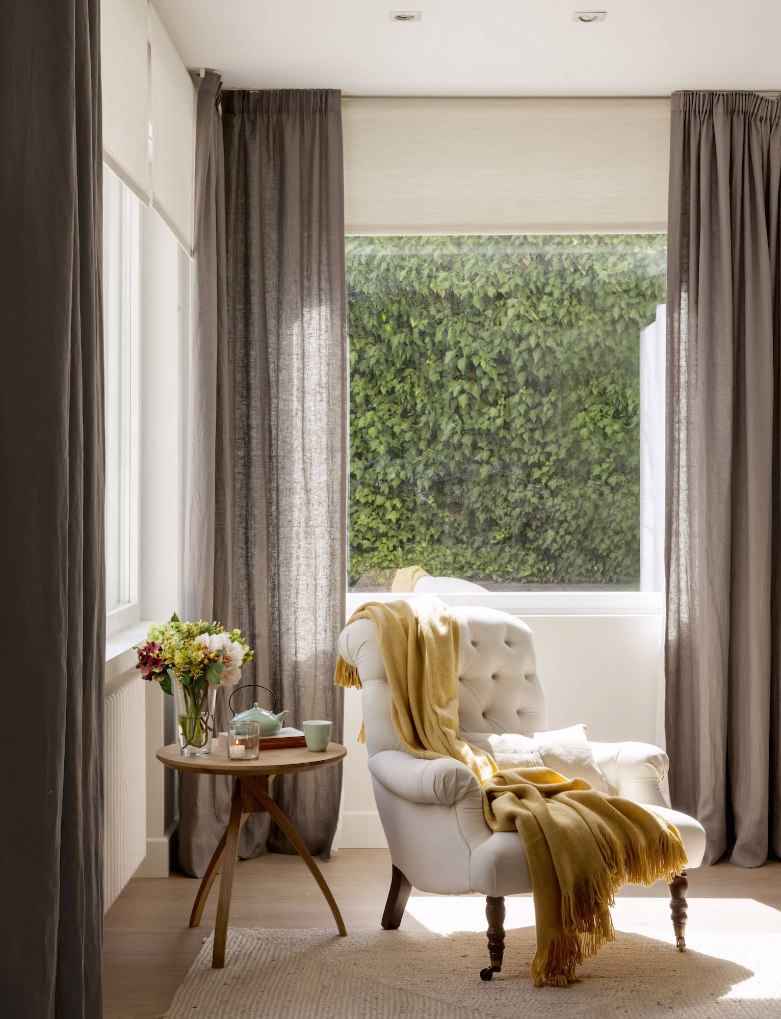 Rincón del salón con butaca blanca y ventanas con cortinas grises. 