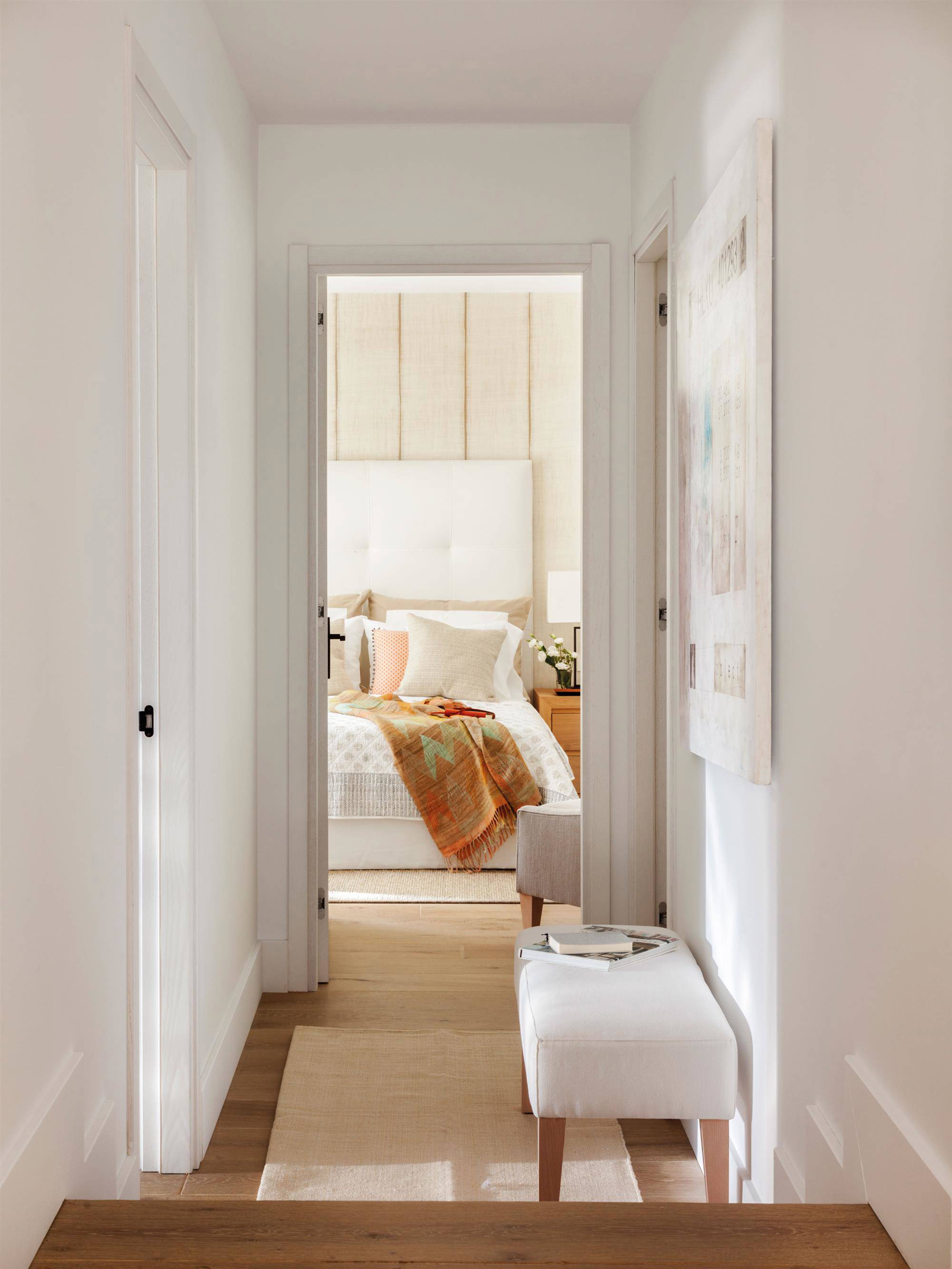 Pasillo con escalera, alfombra, taburete y cuadro. Al fondo, el dormitorio.