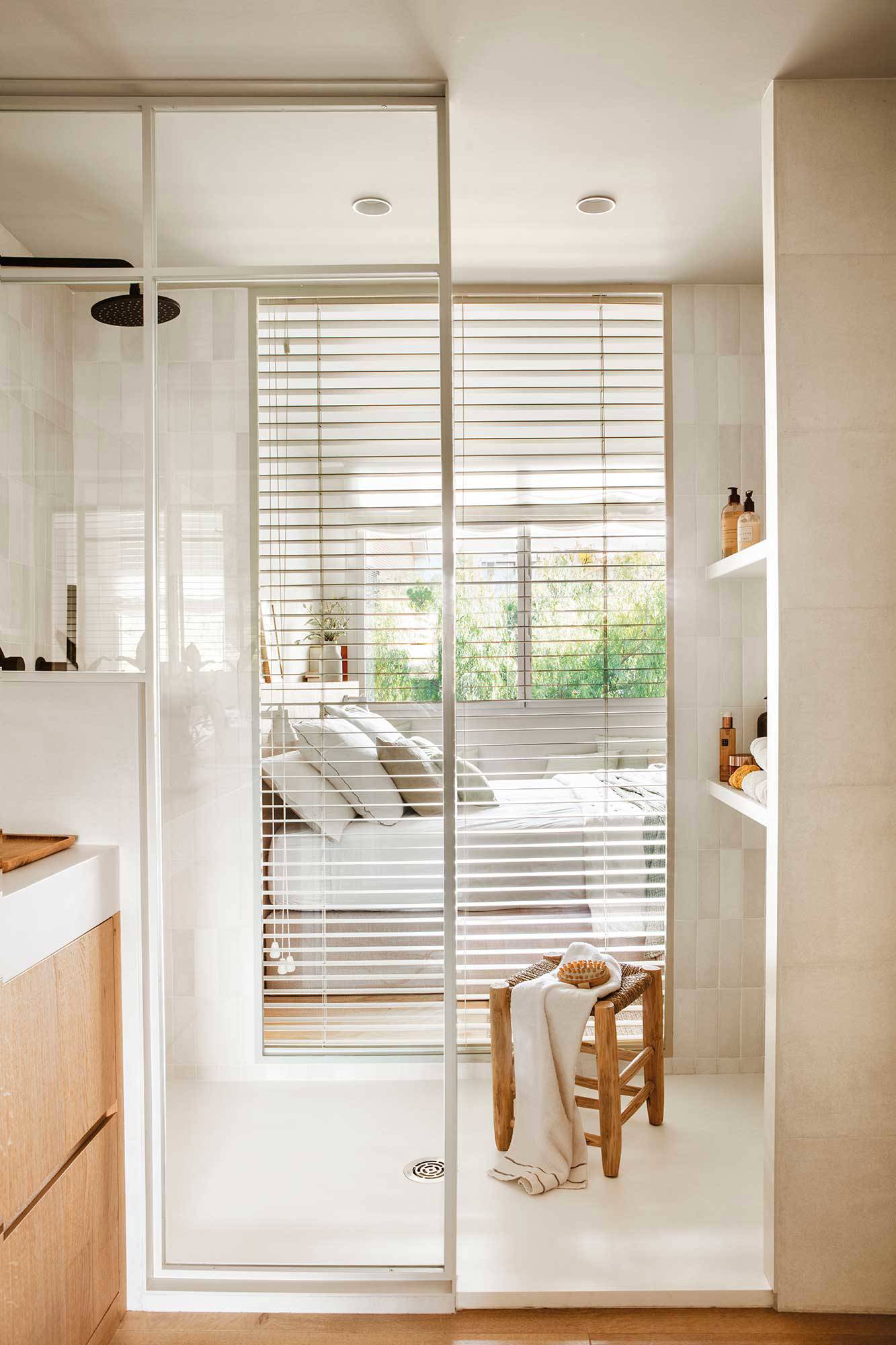 Baño con ducha integrado en el dormitorio a través de una ventana con persiana veneciana.