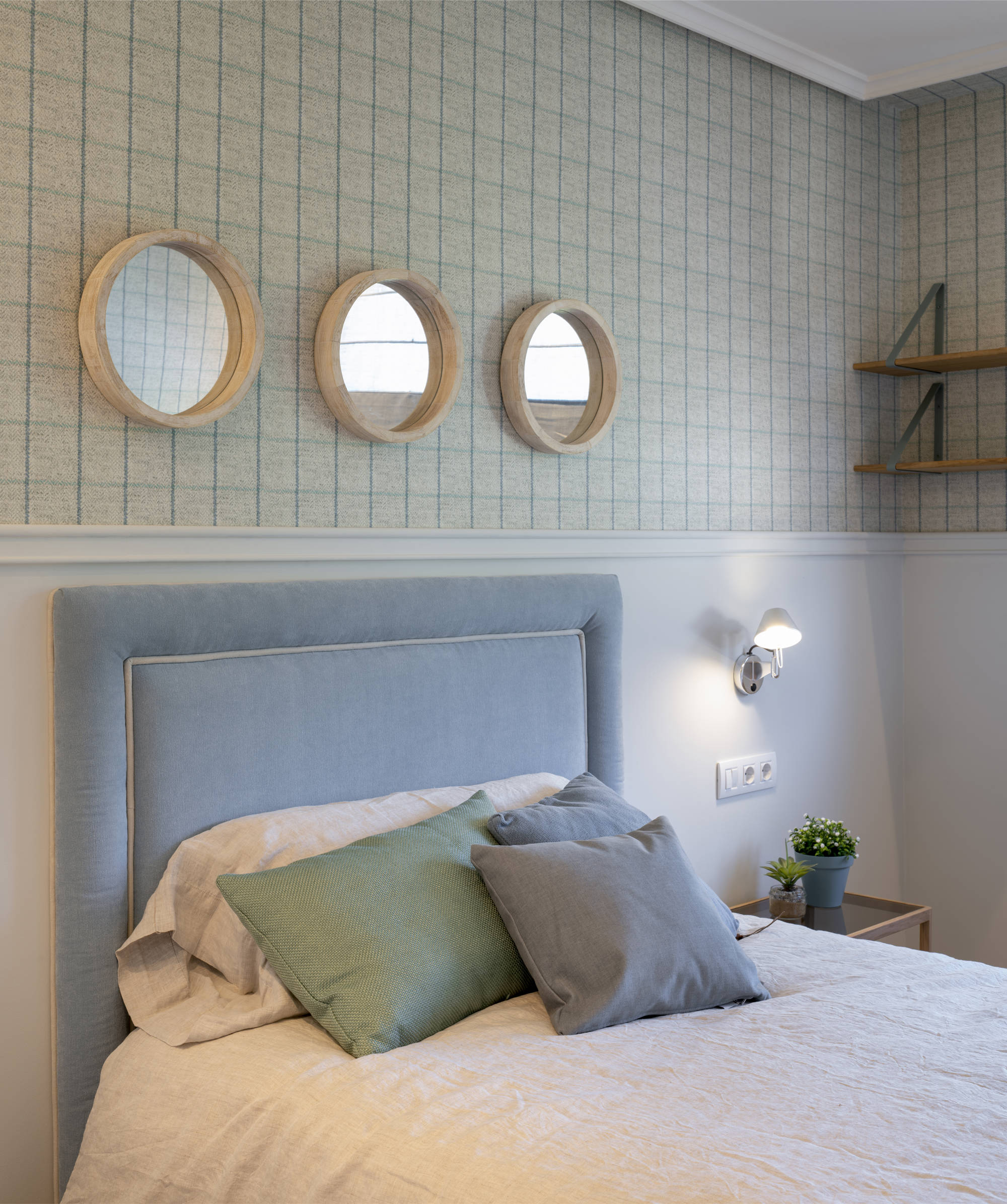 Dormitorio con tres espejos redondos, zócalo y papel pintado y cama con cabecero tapizado en azul