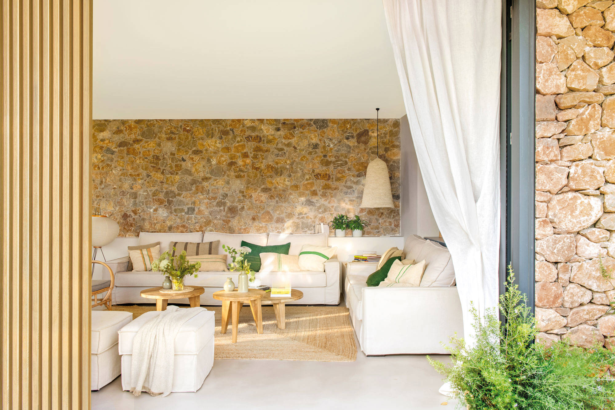 Salón de primavera con pared de piedra, sofás blancos y textiles en tonos tierra y verdes.