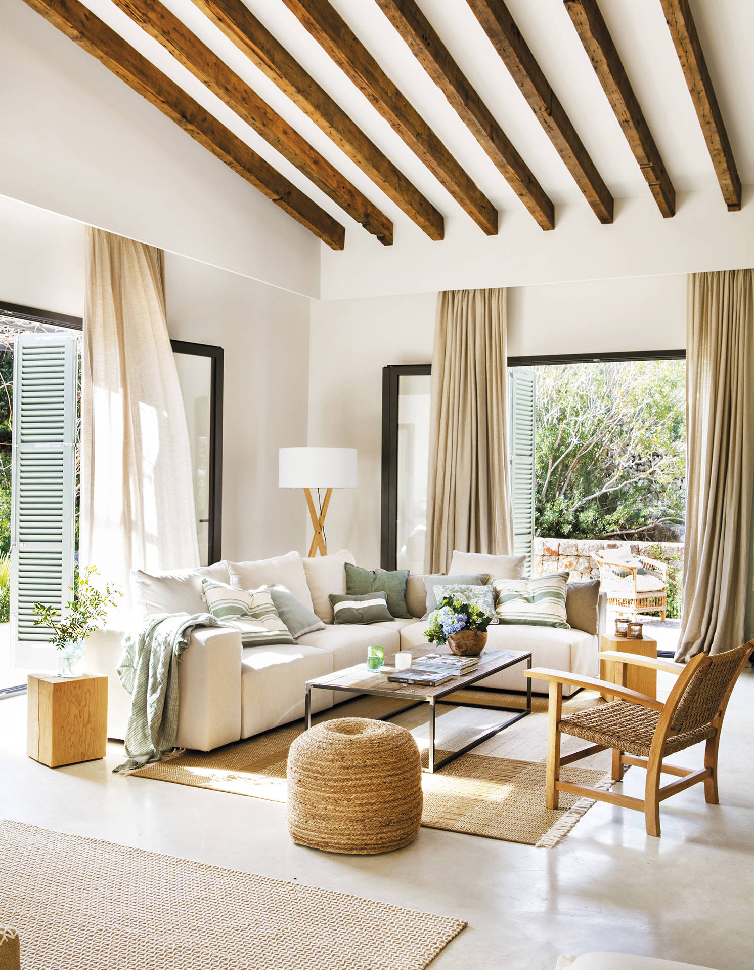 Salón de verano con sofá rinconera blanco, puf y alfombra de yute, butaca de madera y mesa de centro de madera y metal.