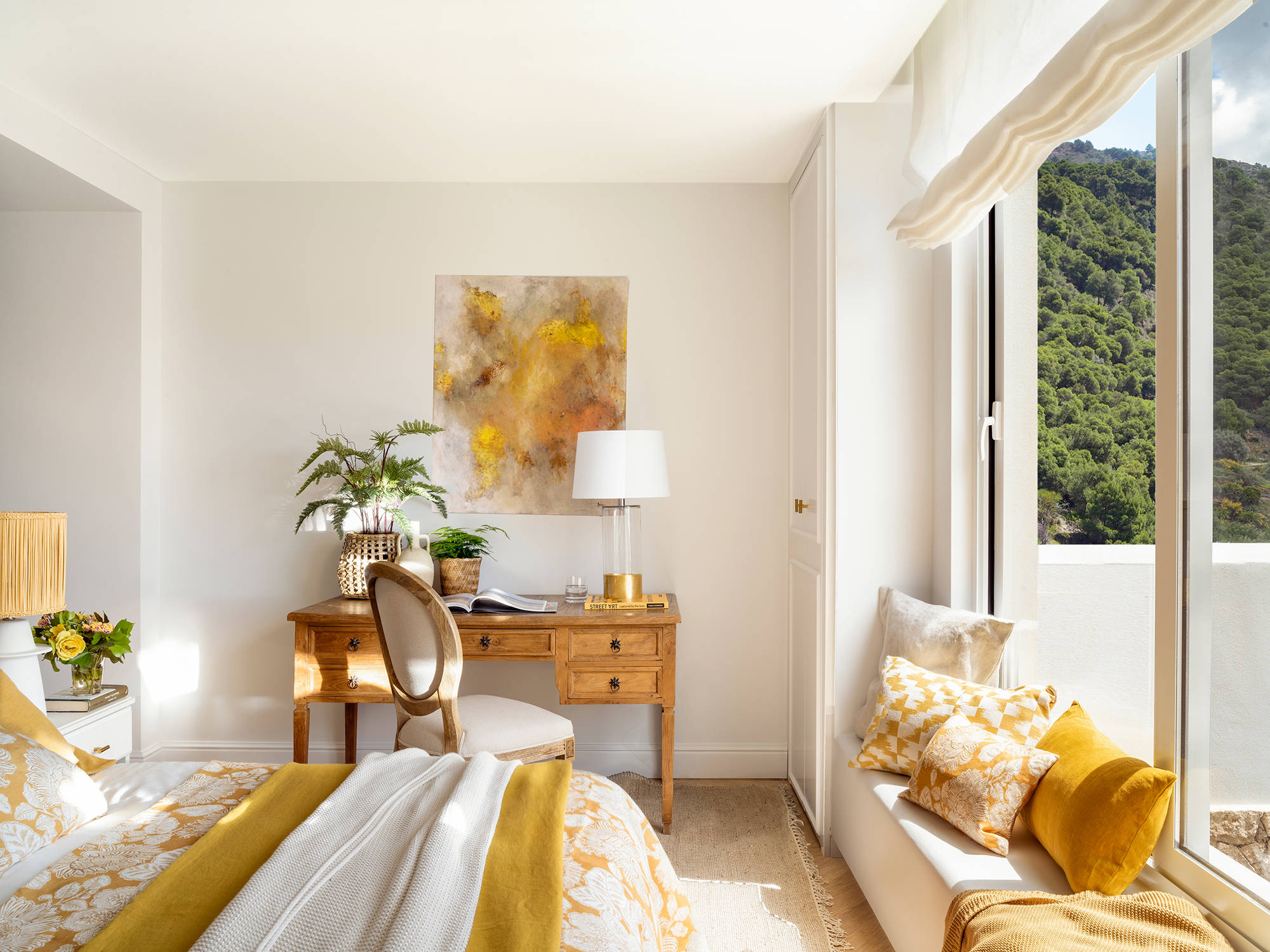 Dormitorio con escritorio de madera, cuadro y banco bajo la ventana con cojines color mostaza.