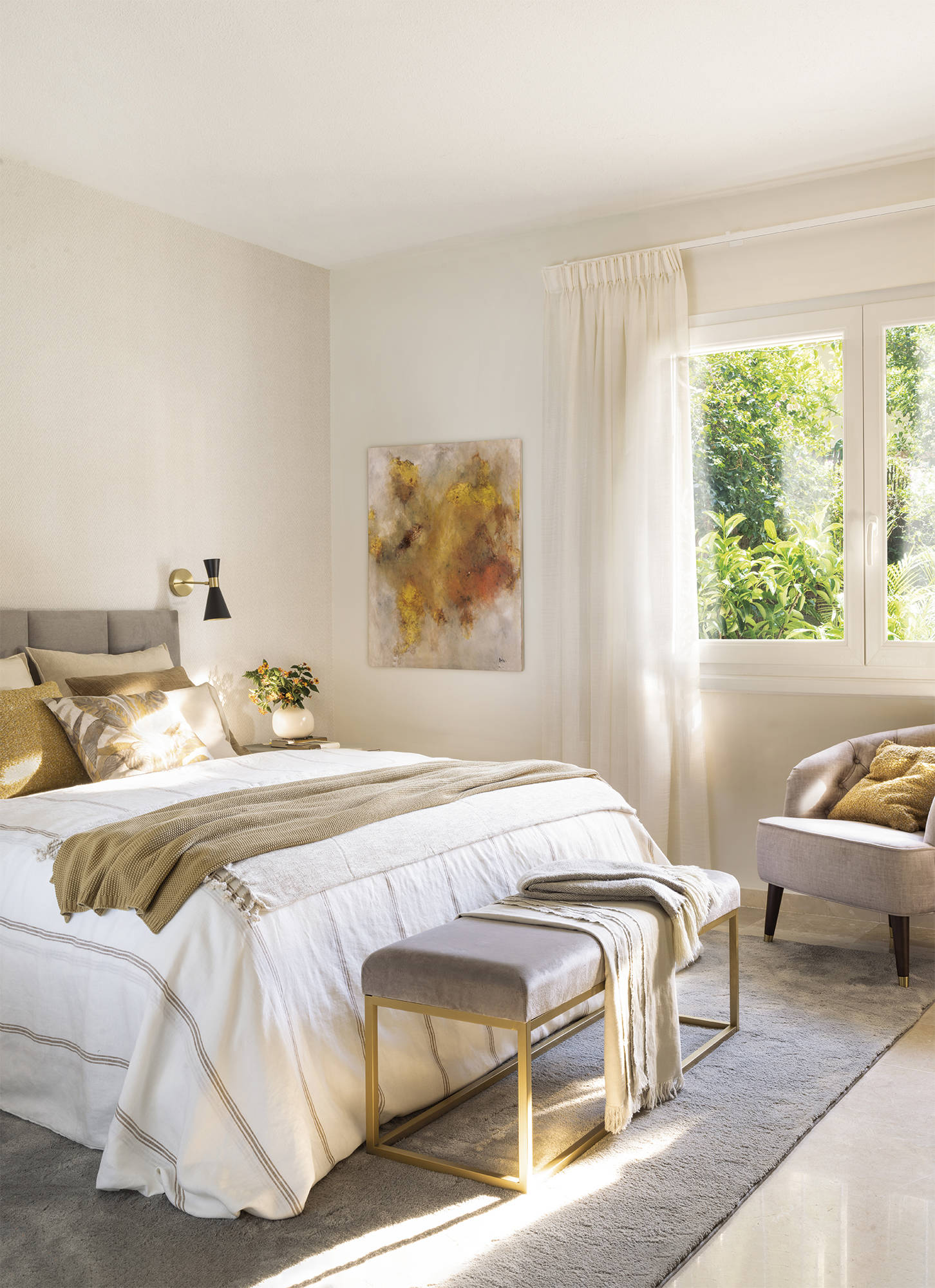 Dormitorio con cabecero tapizado, aplique dorado, banqueta, butaca tapizada y cuadro moderno