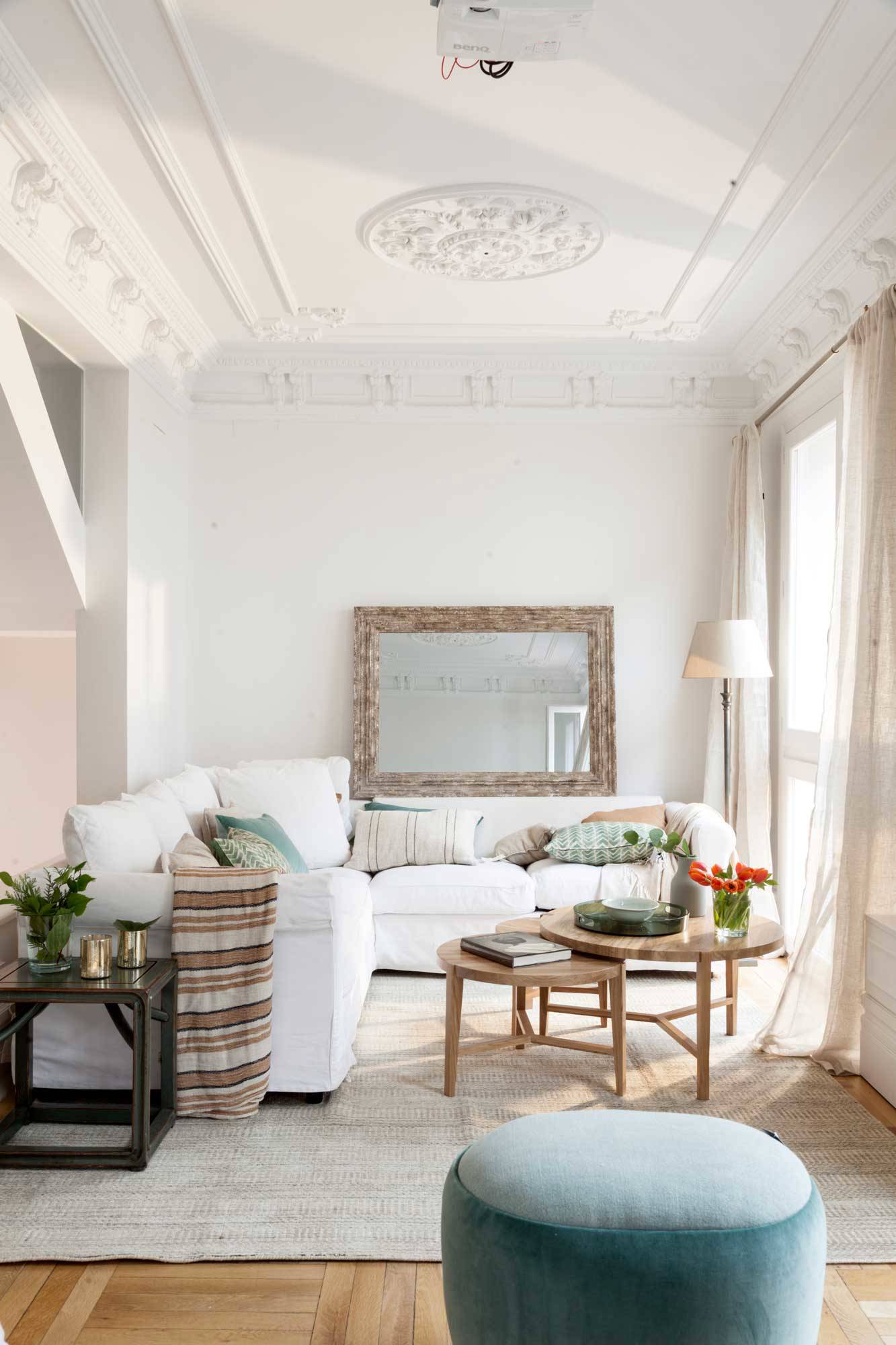 Salón luminoso en blanco con molduras en el techo y espejo sobre el sofá.