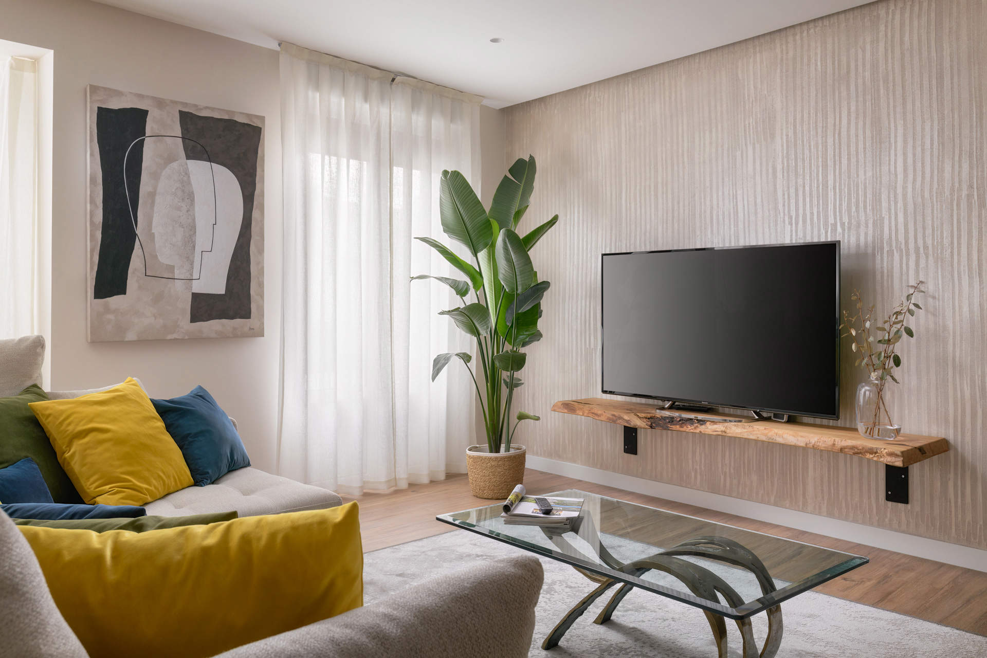 Salón con sofá gris, mesa de centro con sobre de cristal, cuadro, planta de interior y mueble de TV de madera en forma de balda suspendida.
