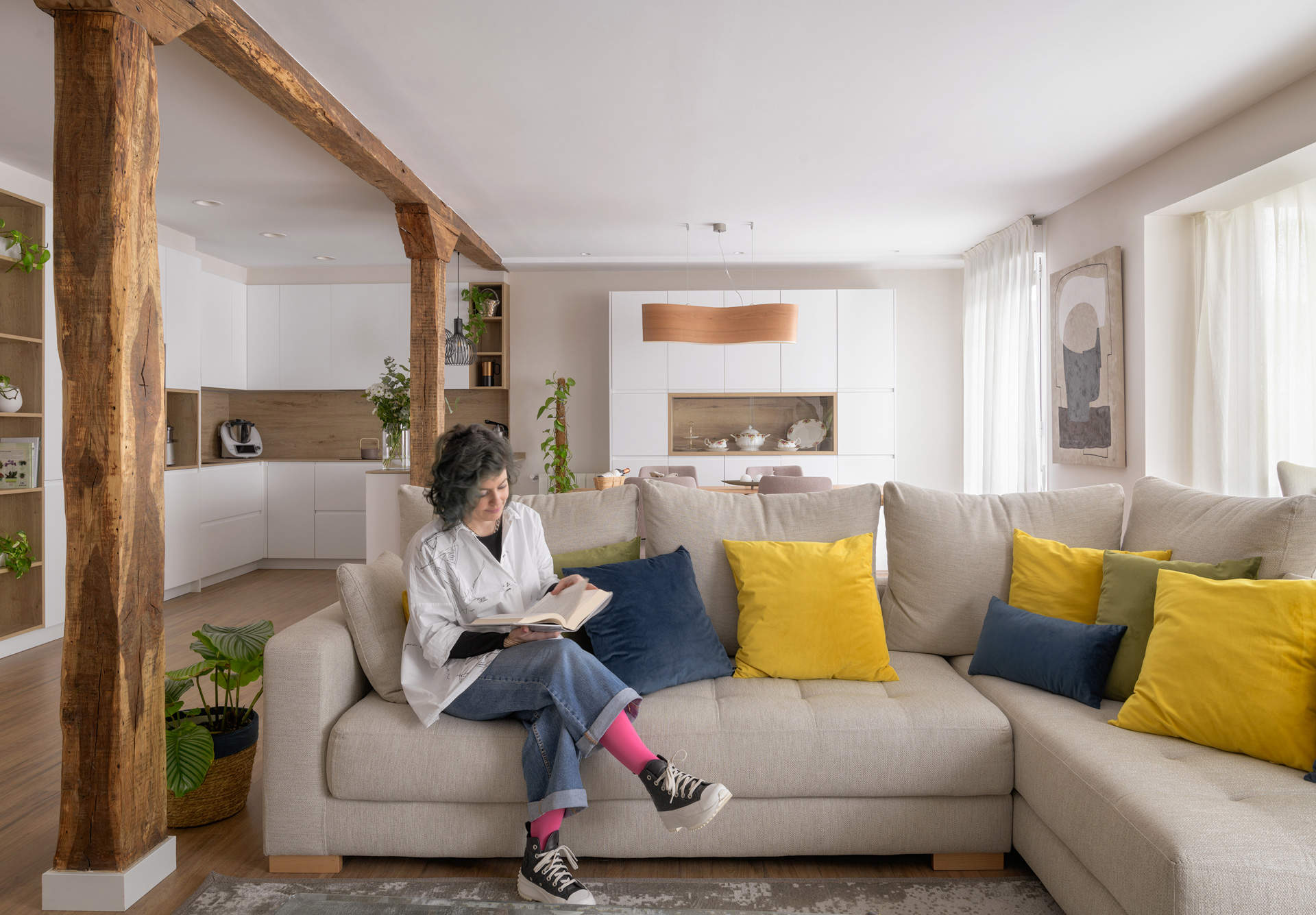 Salón con sofá gris en forma de L, cojines de colores, vigas de madera y cocina al fondo. 