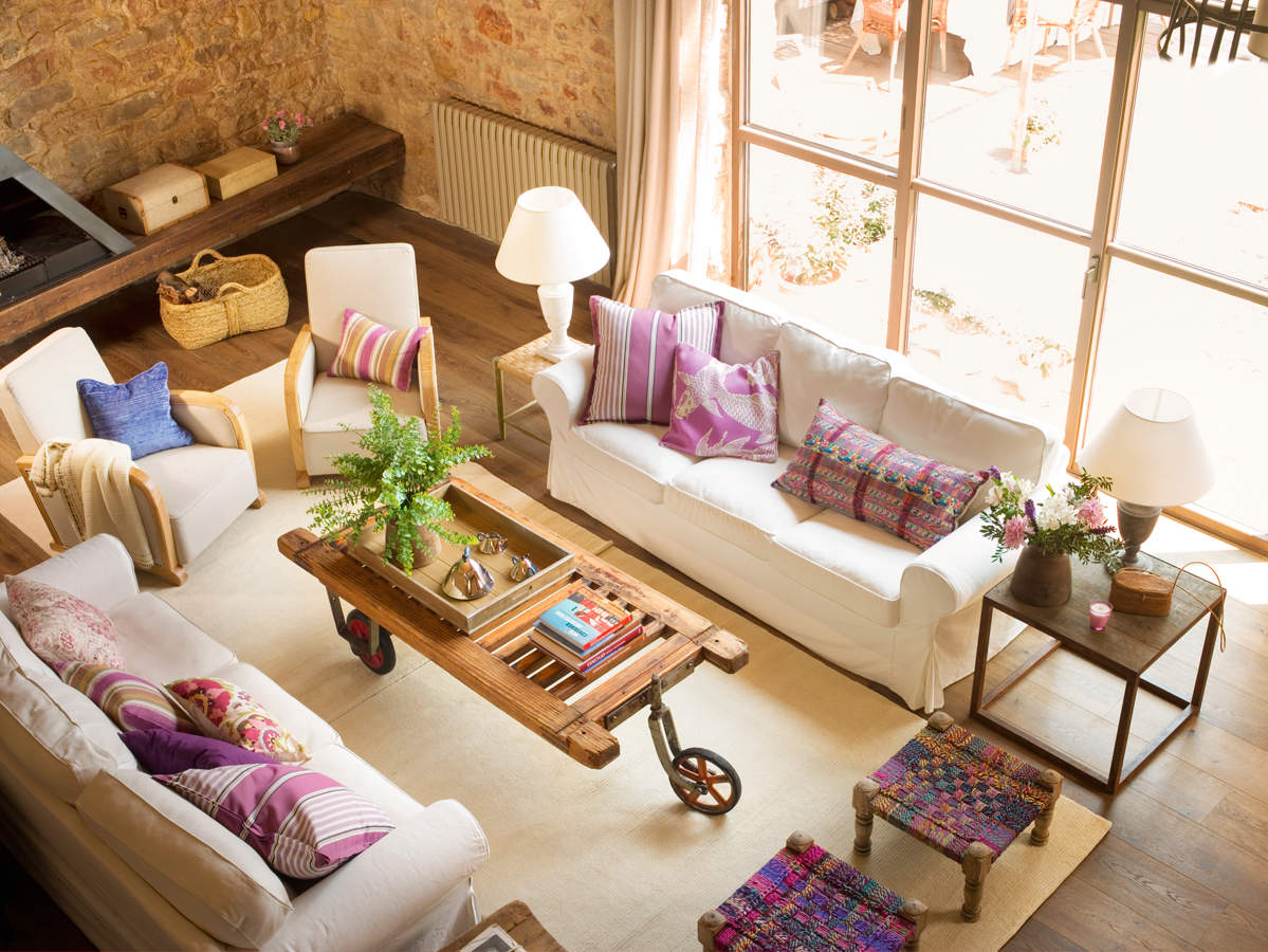 Salón con sofá blanco, banquetas artesanas en madera y tejido de colores, mesa de centro antigua con ruedas y alfombra.