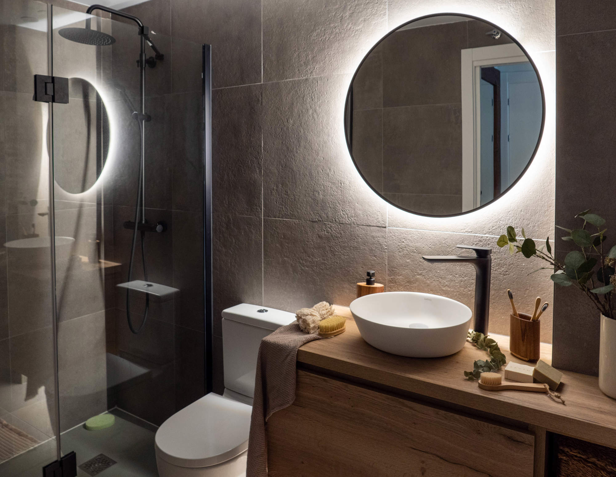 Baño con espejo redondo iluminado, ducha y mueble de madera.