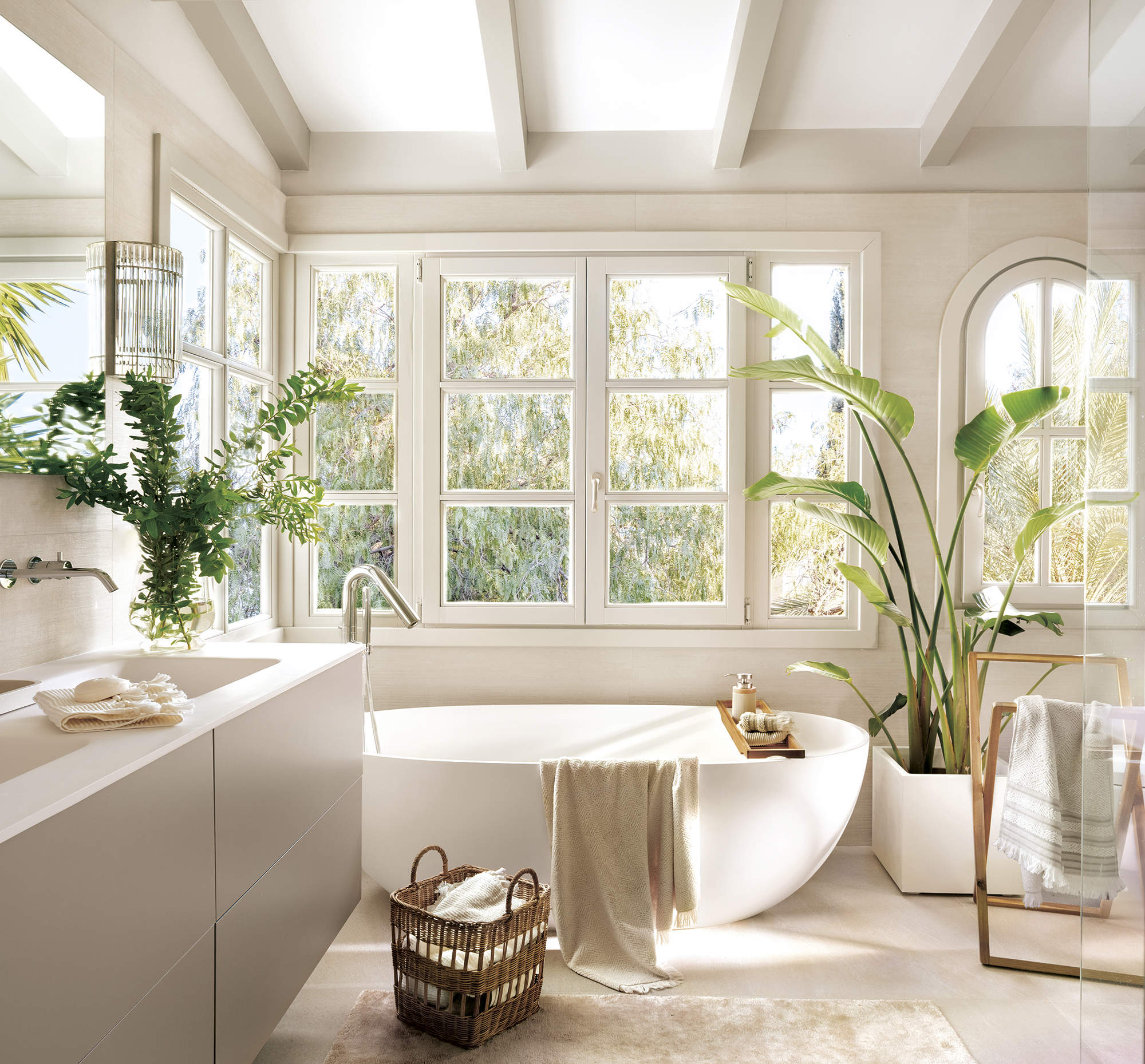baño grande con bañera externa blanca y mueble de lavabo de dos senos, plantas, suelo de piedra con alfombra beige, gran ventanal