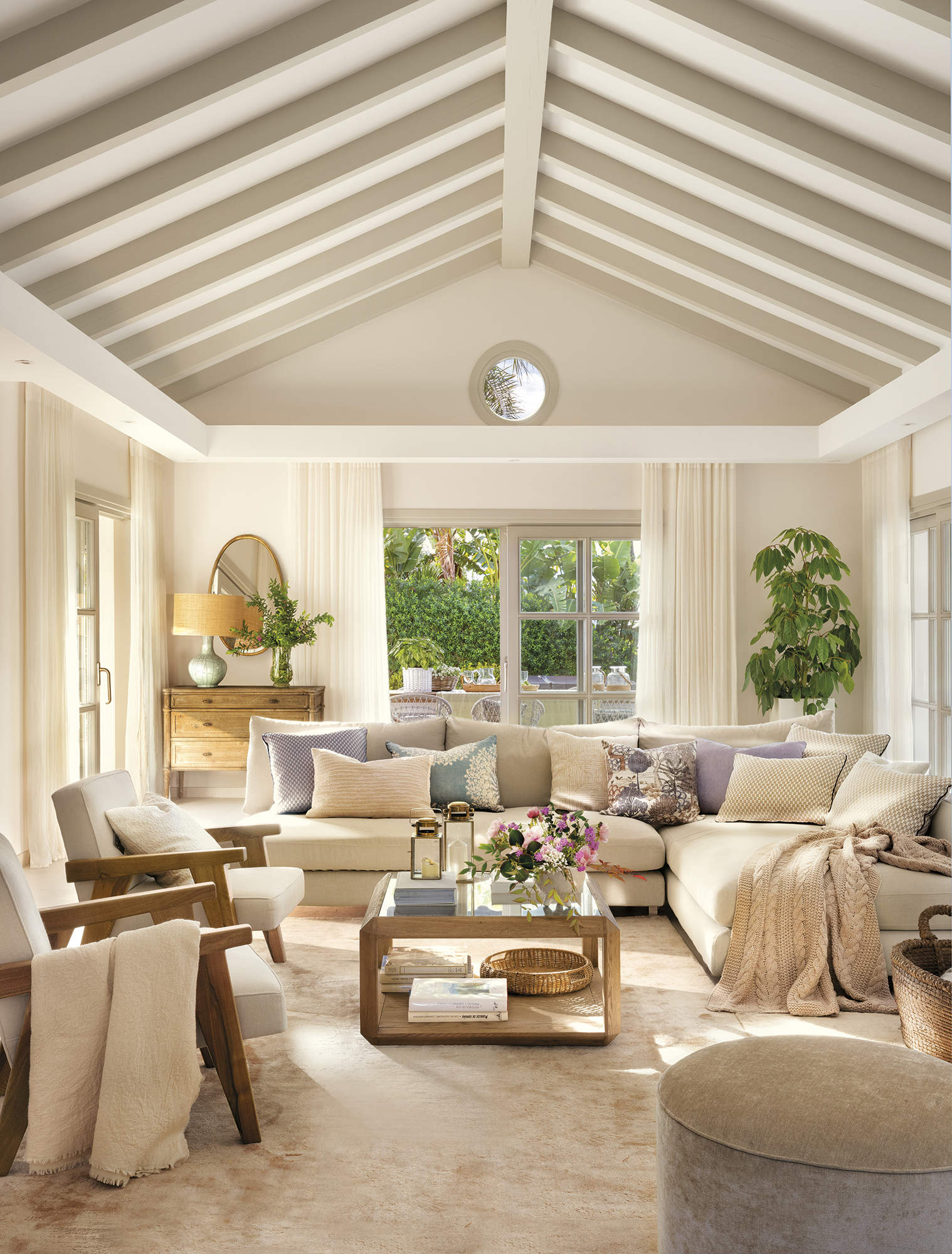 salón blanco con techo a dos aguas con vigas de madera beige, sofá esquinero, butacas de madera tapizadas, alfombra beige, puf