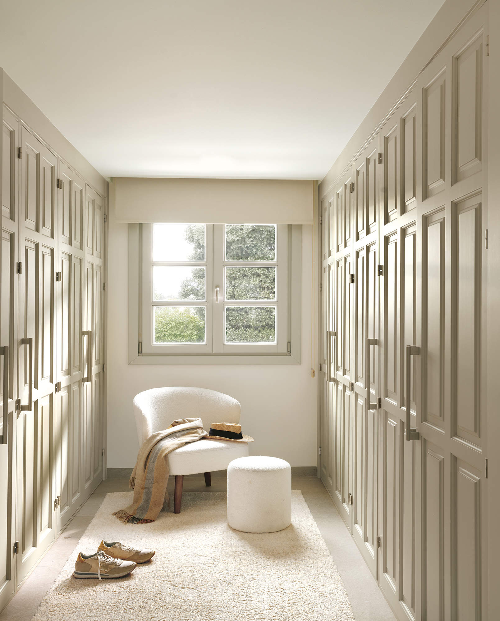 vestidor con armarios enfrentados de madera pintada de beige, suelo de piedra, alfombra beige con butaca y puf tapizados, ventana con estor