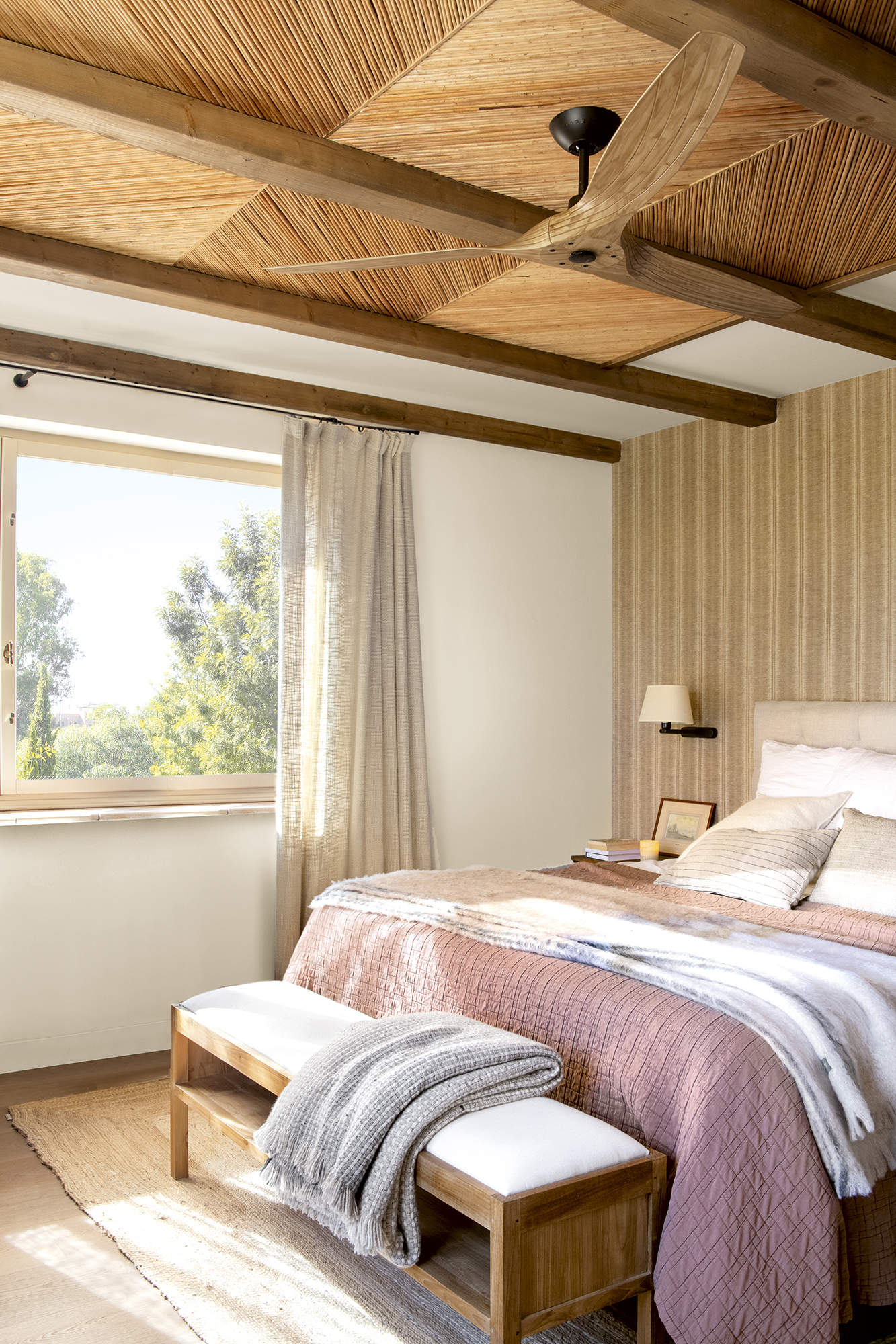Dormitorio con pared empapelada y techo de fibra, ropa de cama rosa y banqueta blanca.