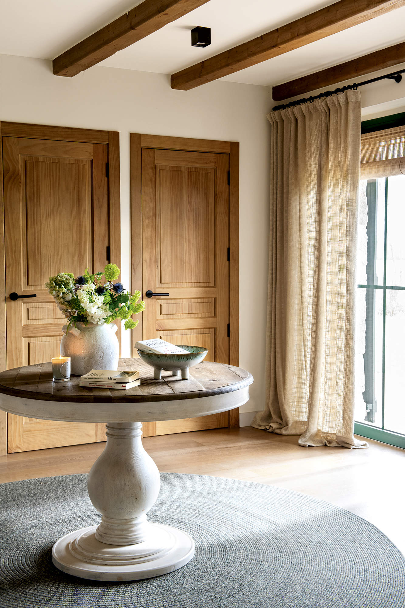 Recibidor con mesa redonda de madera con alfombra redonda gris, suelo de madera, puertas de madera y vigas