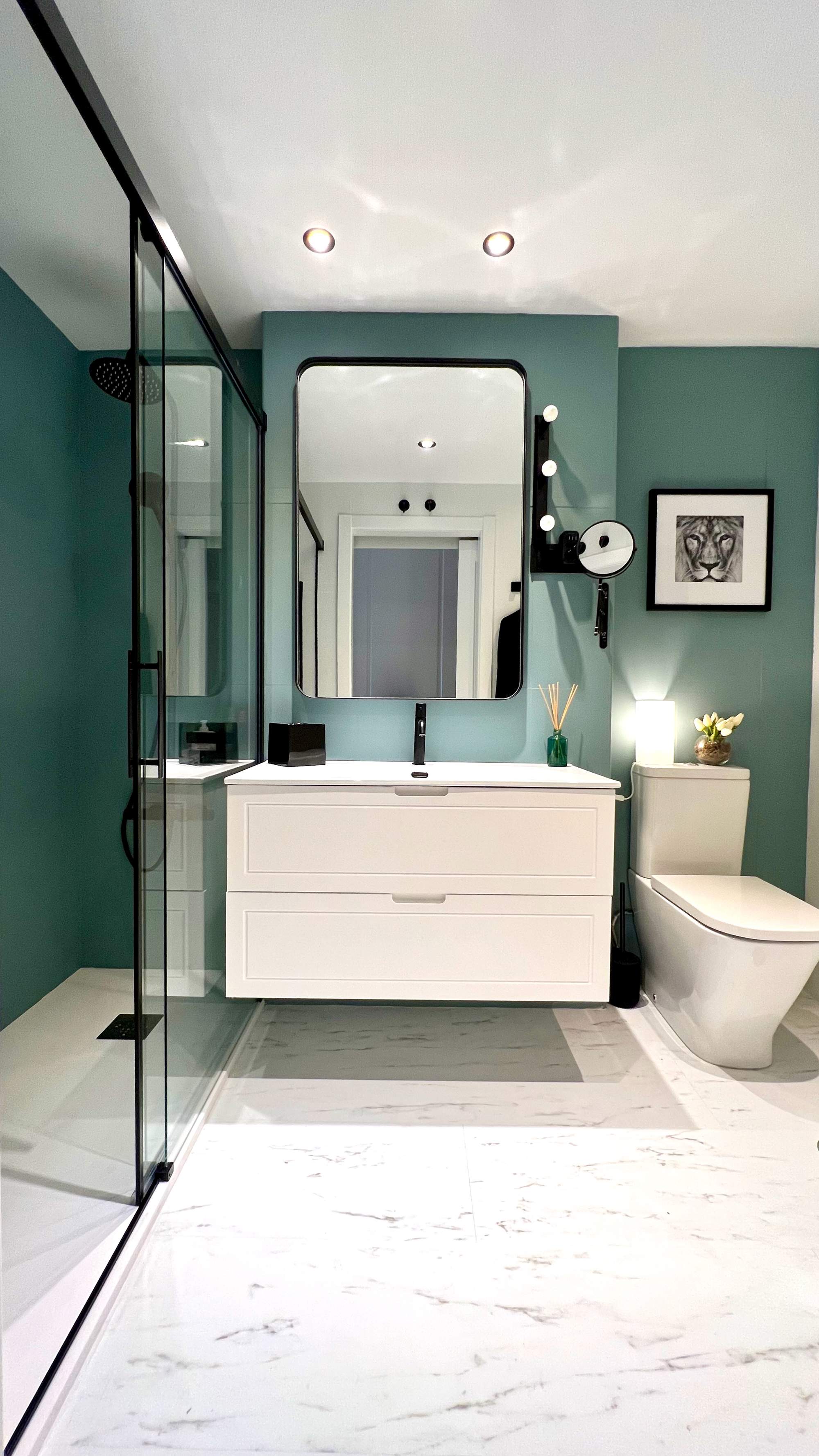 Baño con pared pintada en verde, mueble bajolavabo lacado en blanco y ducha con mampara. Pavimento, de suelo vinílico que imita el mármol.
